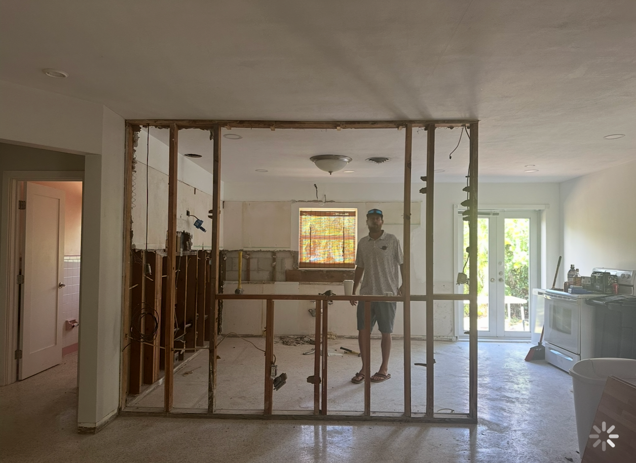 Man working on a home renovation in a room with walls partially demolished, exposing wall studs and electrical wiring, and construction tools scattered around.