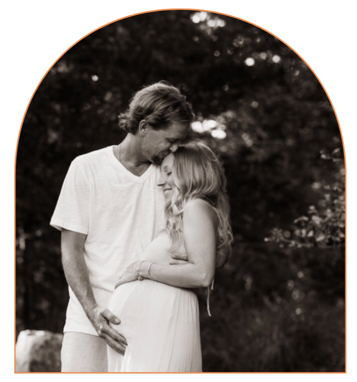 A couple share a tender moment outdoors, with the man gently holding the woman's pregnant belly and touching foreheads, both smiling with eyes closed.