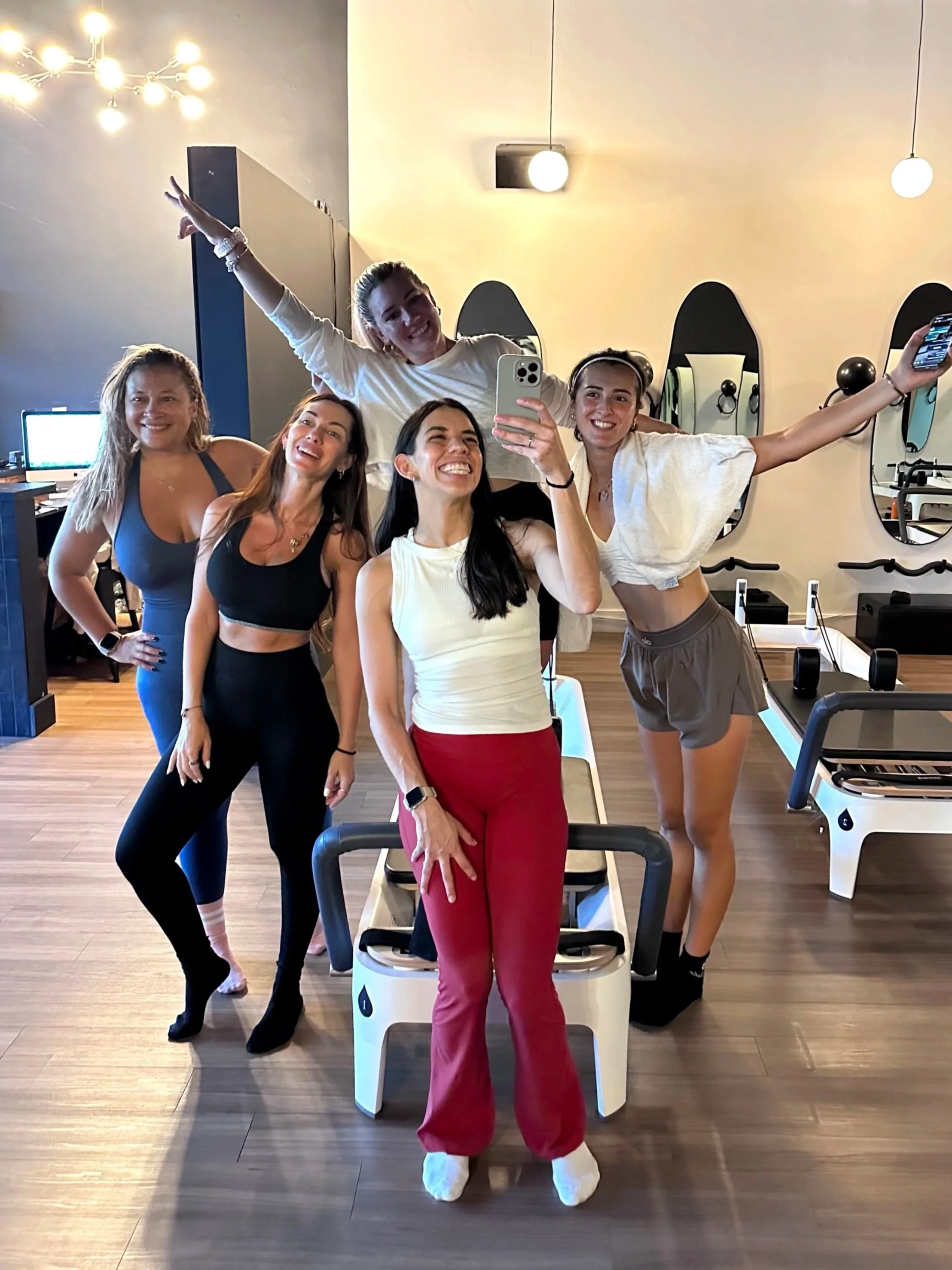 Six women in athletic clothing posing for a selfie in a gym or fitness studio, with exercise equipment and mirrors in the background.
