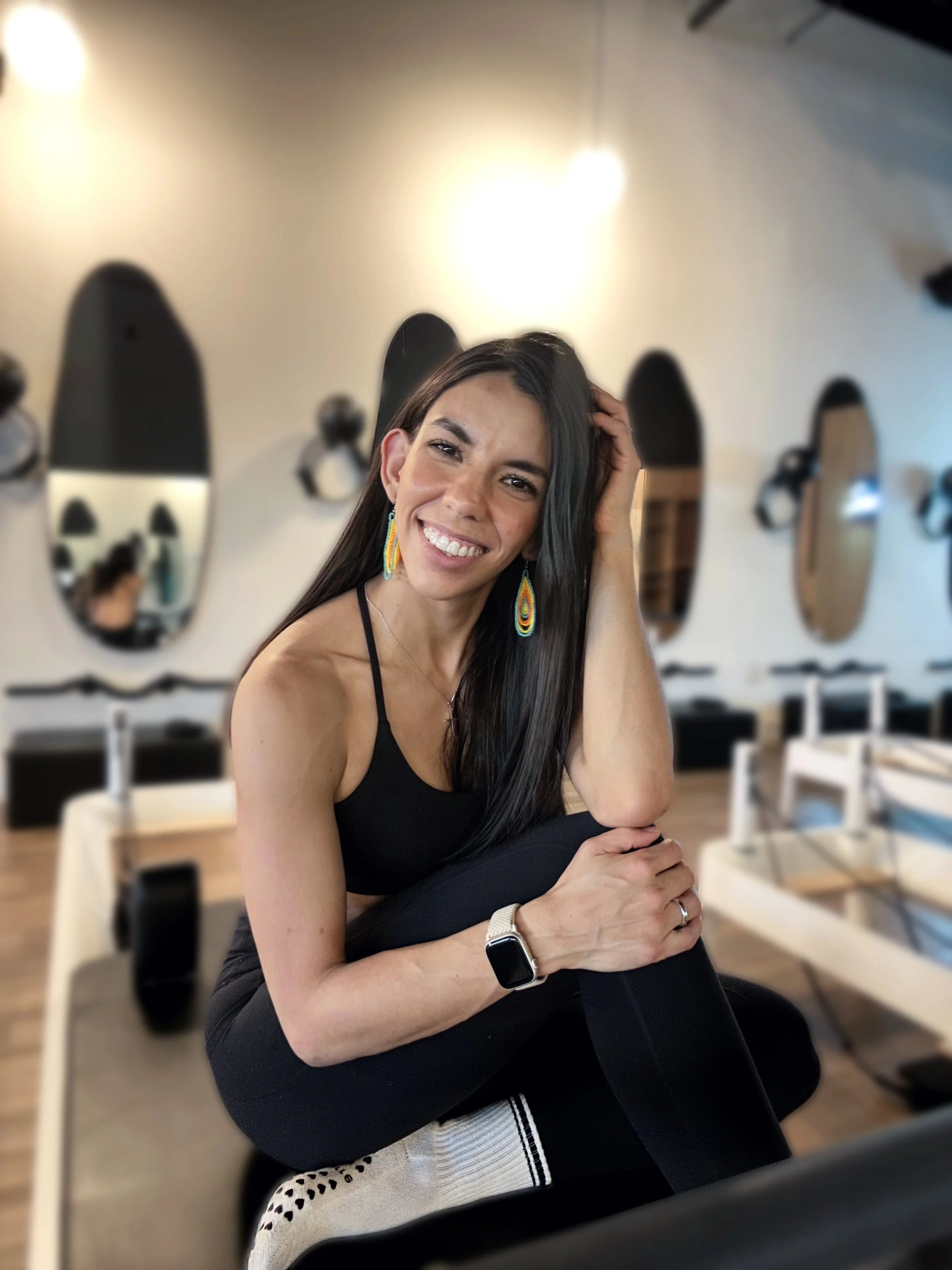 Young woman with dark hair, colorful earrings, smiling, sitting in a restaurant with mirrors on the wall, wearing a black top and a smartwatch.