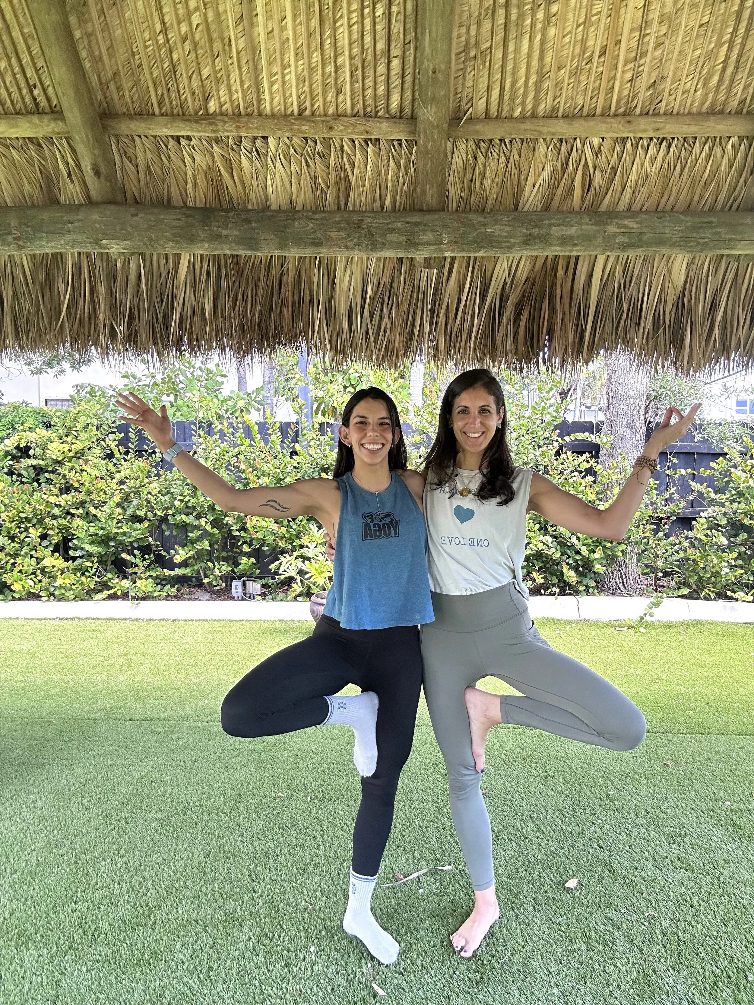 Two women smiling and practicing yoga together outdoors under a thatched roof, standing on one leg with arms raised and one foot placed against the inner thigh of the opposite leg, in a yoga pose.