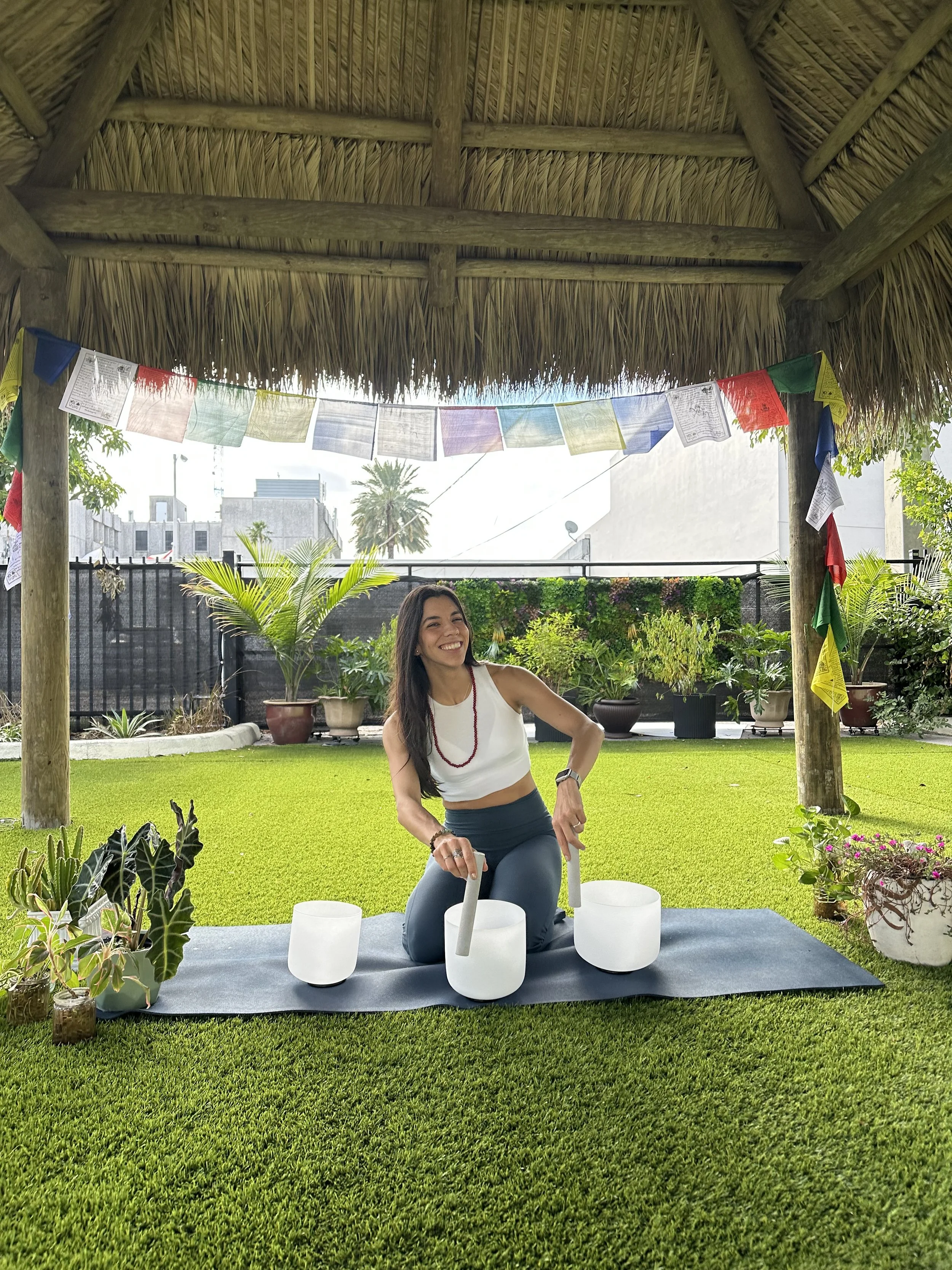 A woman practicing sound therapy with singing bowls outdoors under a thatched roof structure.
