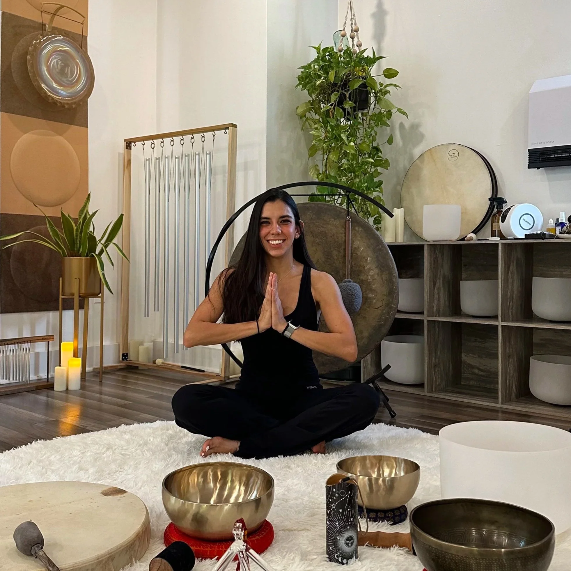 Woman practicing meditation or yoga in a peaceful indoor setting, sitting cross-legged on a white fluffy rug, with metallic singing bowls in the foreground, surrounded by candles, plants, and various spiritual or wellness items.