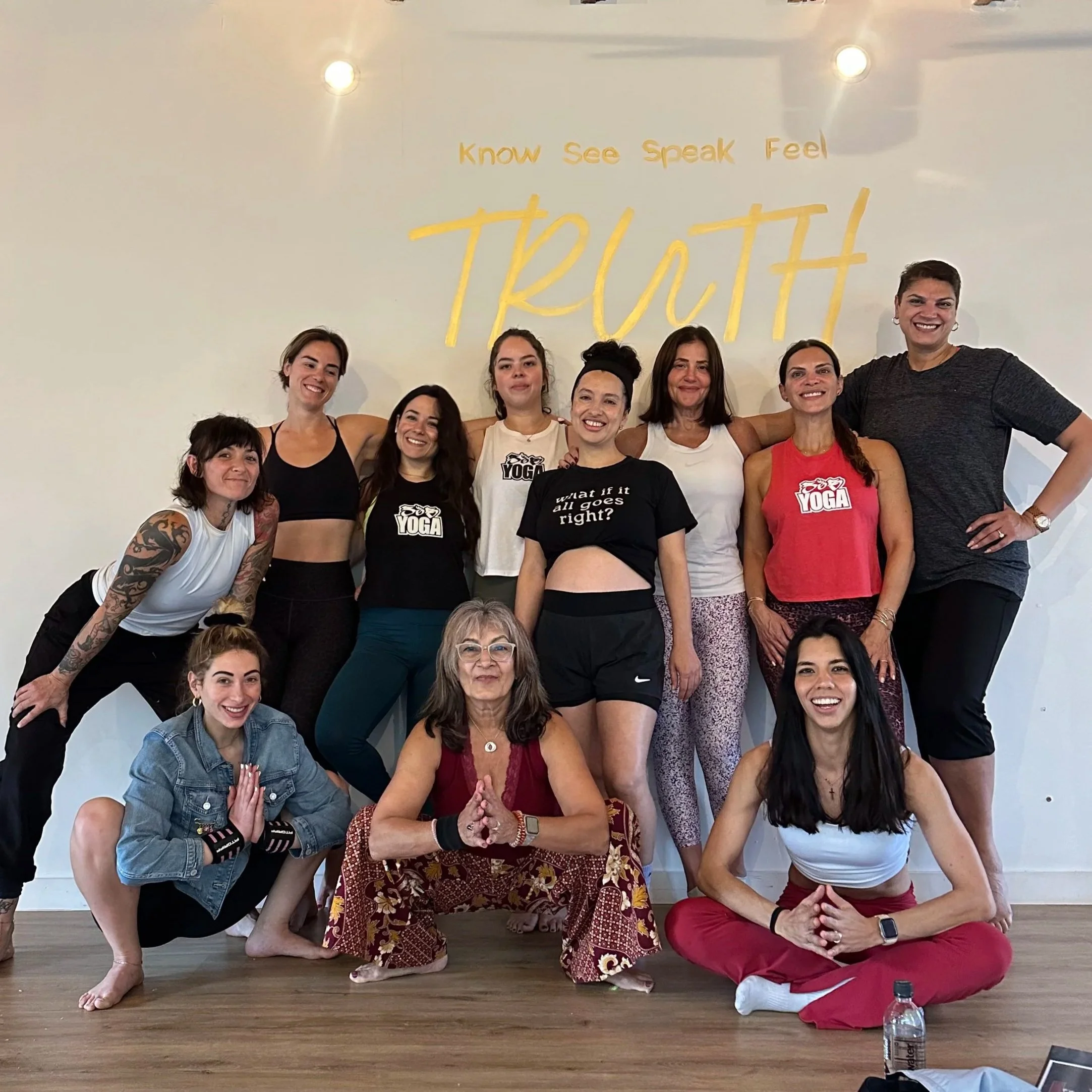 Group of people in a yoga class, some standing and others kneeling or sitting, in front of a white wall with yellow text that reads "know see speak Feel." The group appears to be diverse in age and ethnicity, and they are smiling and relaxed.