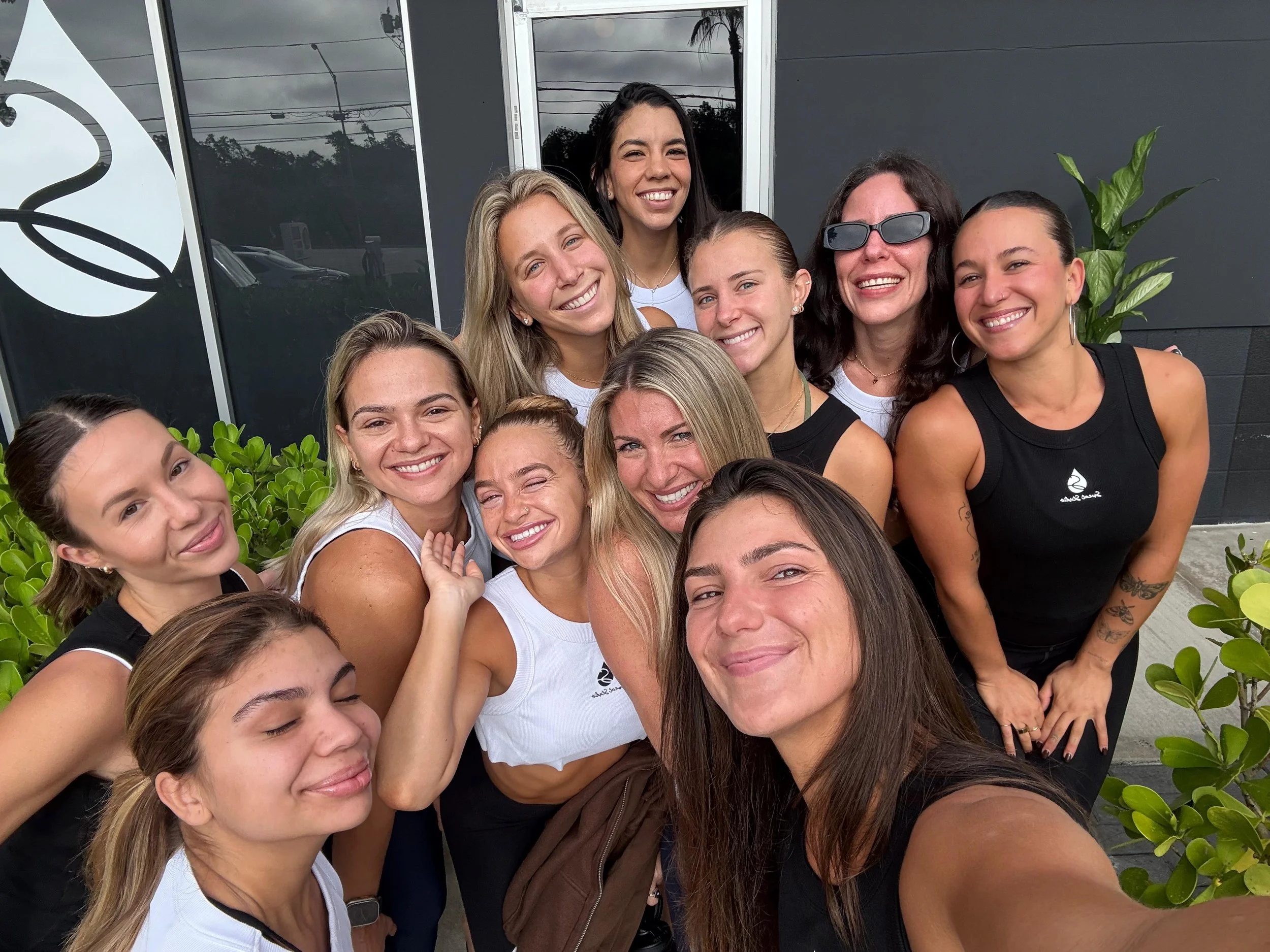 Group of smiling women taking a selfie outdoors against a dark wall, with plants and a window reflecting cloudy sky and power lines.