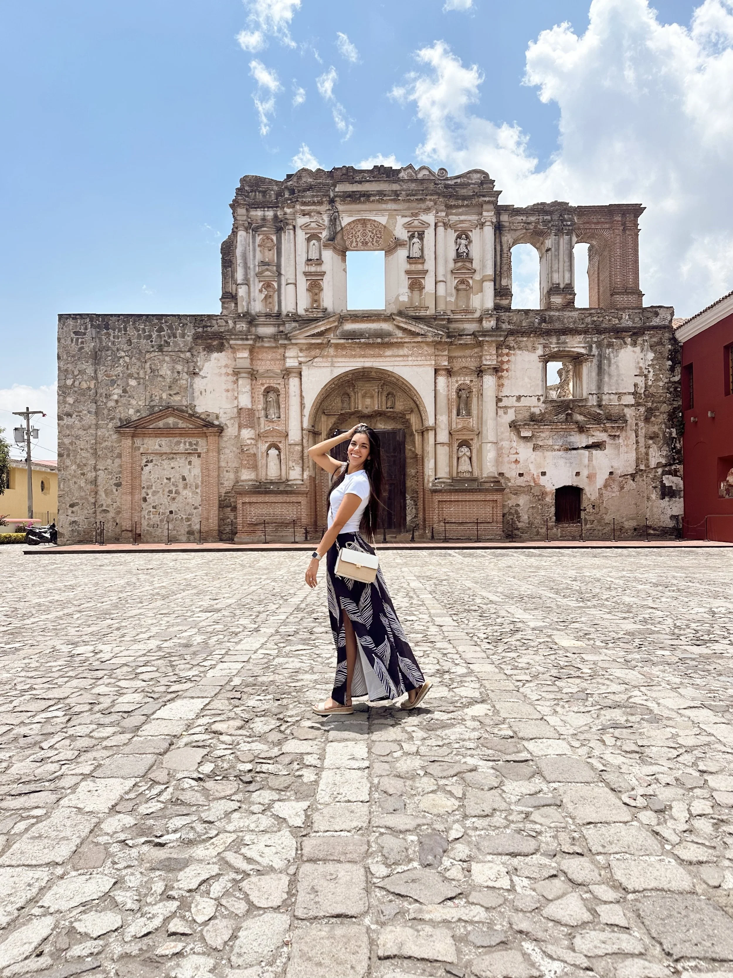 A woman smiling and posing in front of an old, historic stone and brick building with classical architectural details, under a blue sky with scattered clouds.