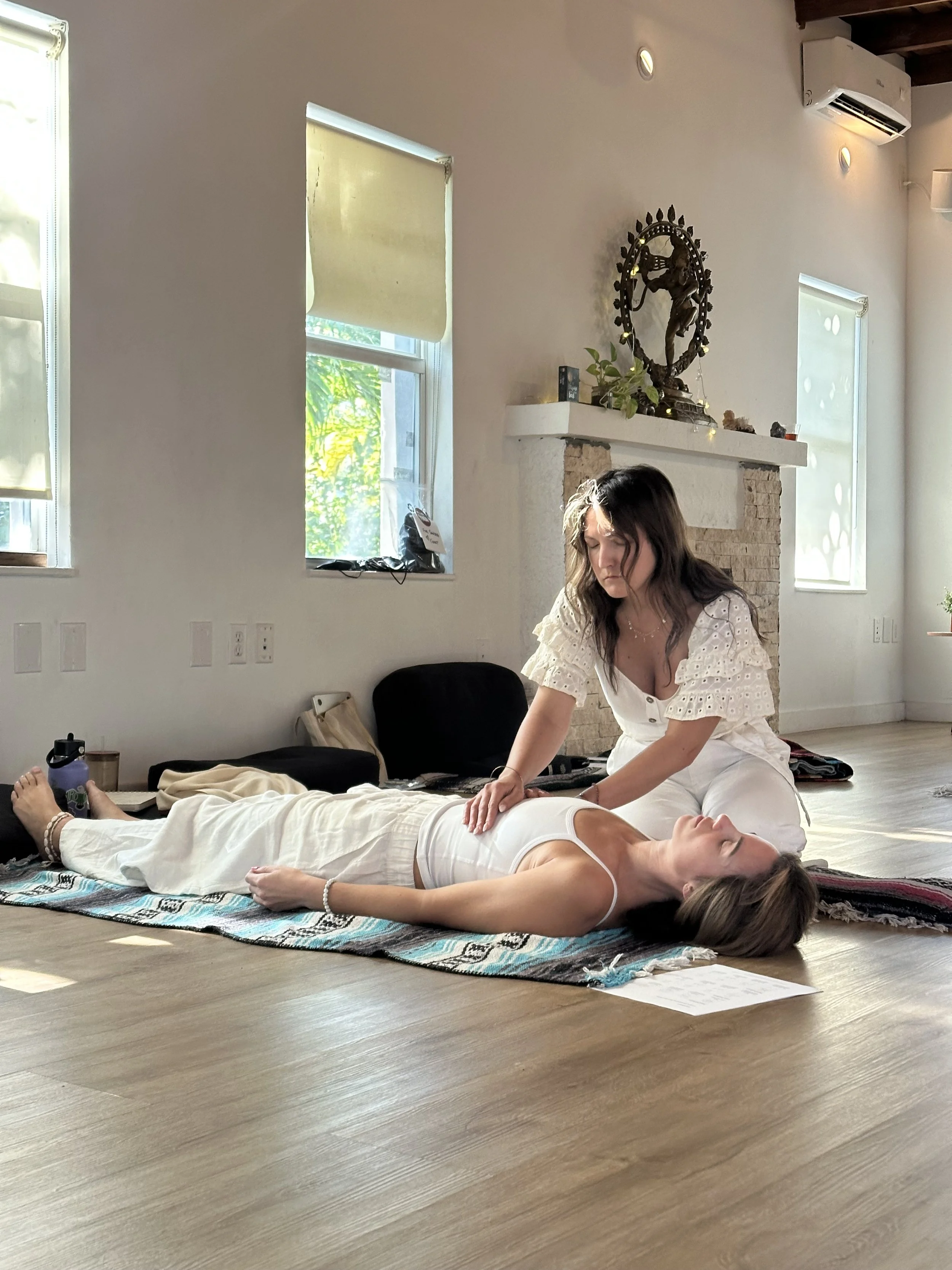 A woman lying on a colorful rug in a room with natural light, receiving a massage from another woman. The room has white walls, two windows, a brick fireplace with a decorative statue and plants, and wooden flooring.