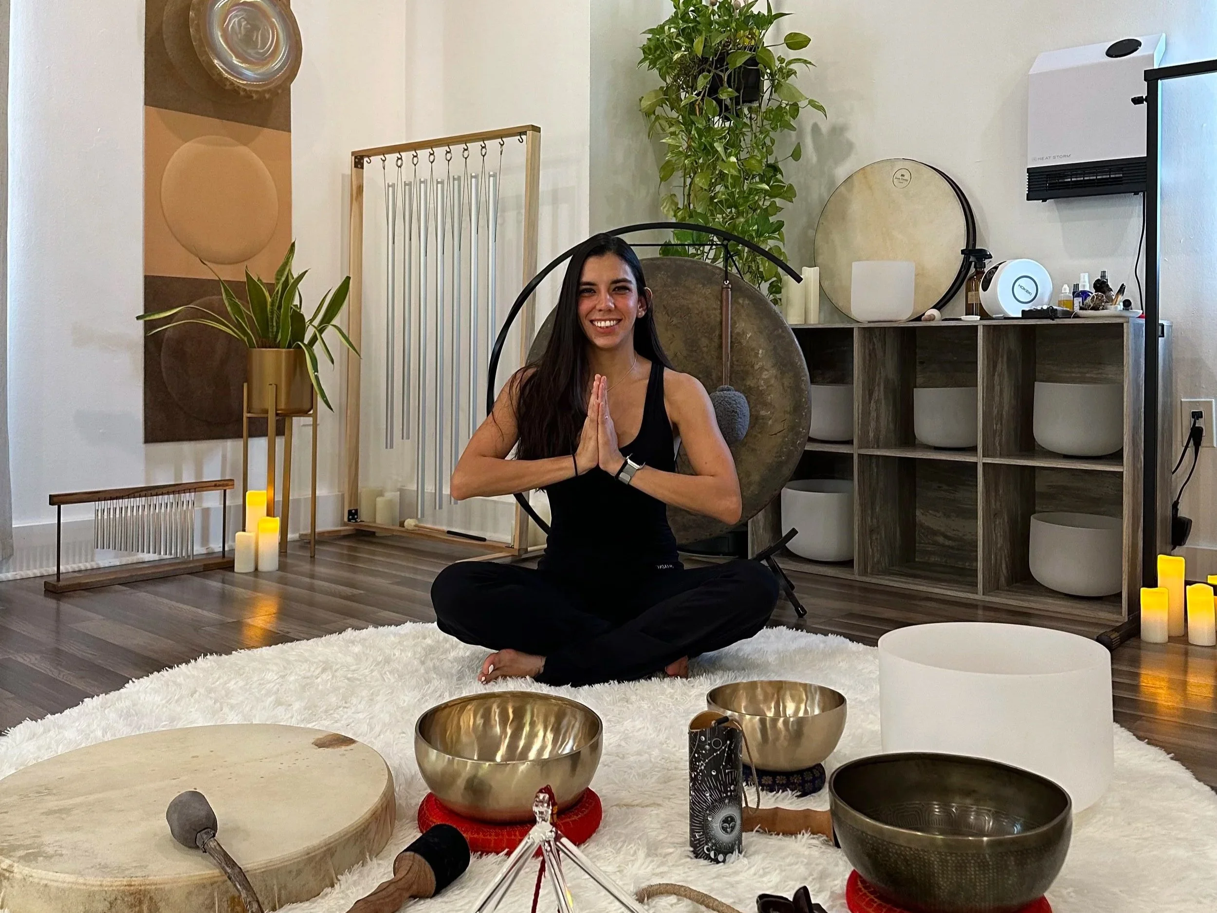 A young woman with long dark hair, sitting cross-legged on a fluffy white rug in a cozy, well-decorated room, smiling and practicing meditation with hands in prayer position. The room has candles, singing bowls, and plants, creating a calming atmosphere.