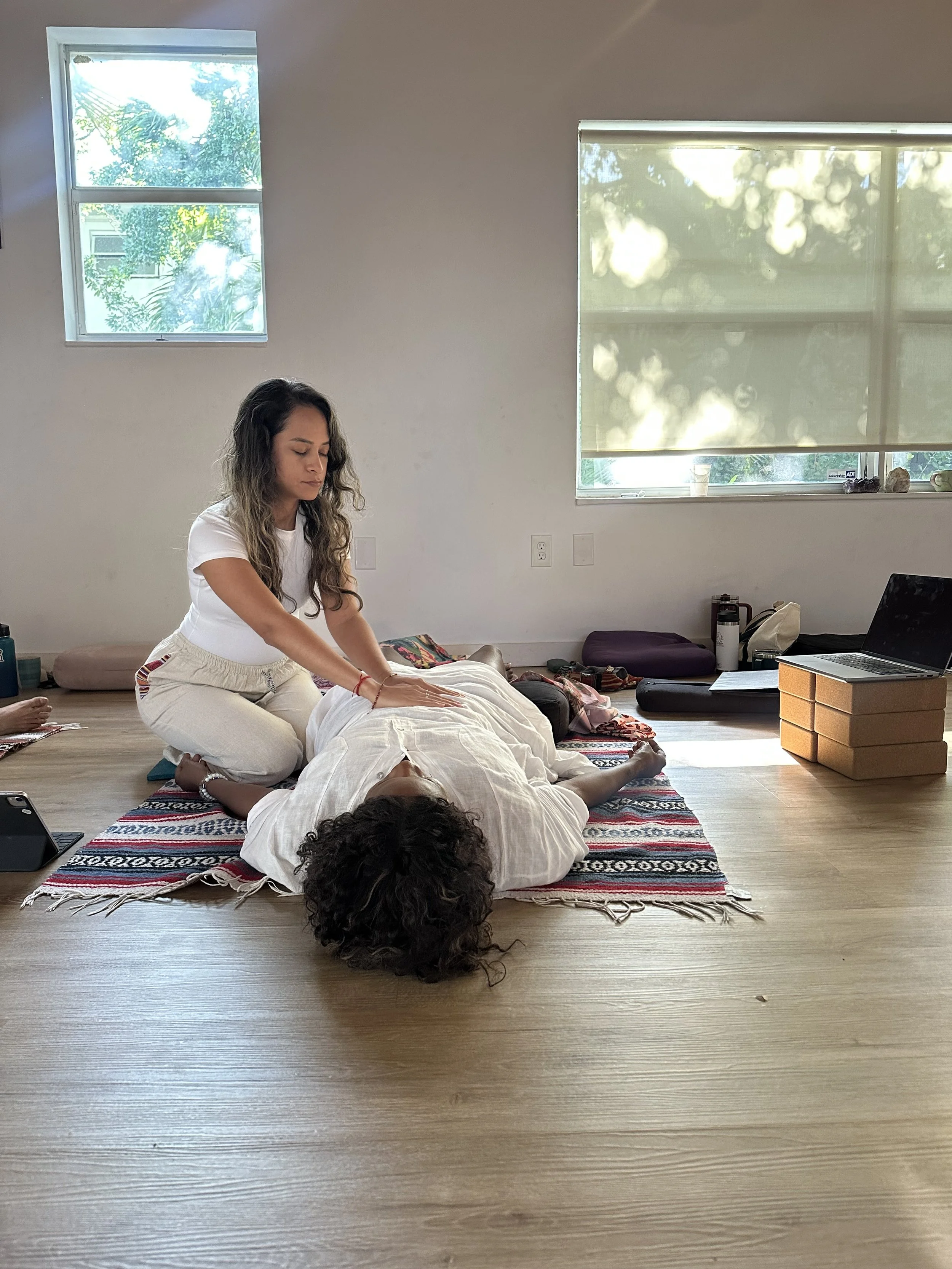 A woman with long wavy hair practicing yoga or stretching on a patterned rug in a bright room with light-colored wooden floors, two large windows with shades, and various items such as a laptop, water bottle, and bags.