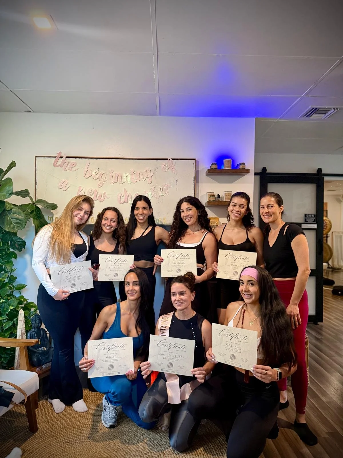 Group of women celebrating with certificates at an indoor event, with a sign that reads 'the beginning of a new chapter' in the background.