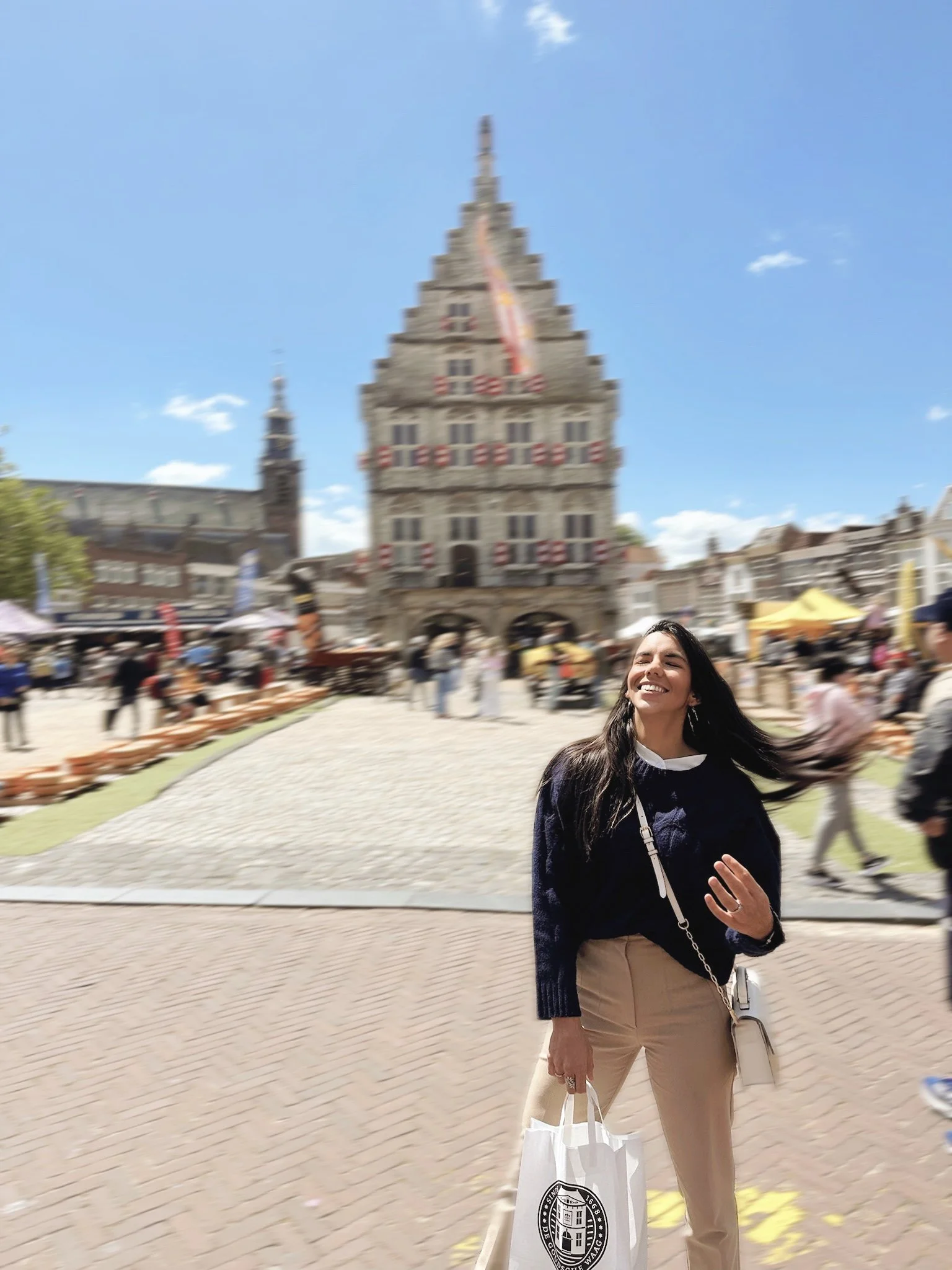 Smiling woman with dark hair wearing a navy sweater and beige pants standing on a busy cobblestone street with historic building in the background.
