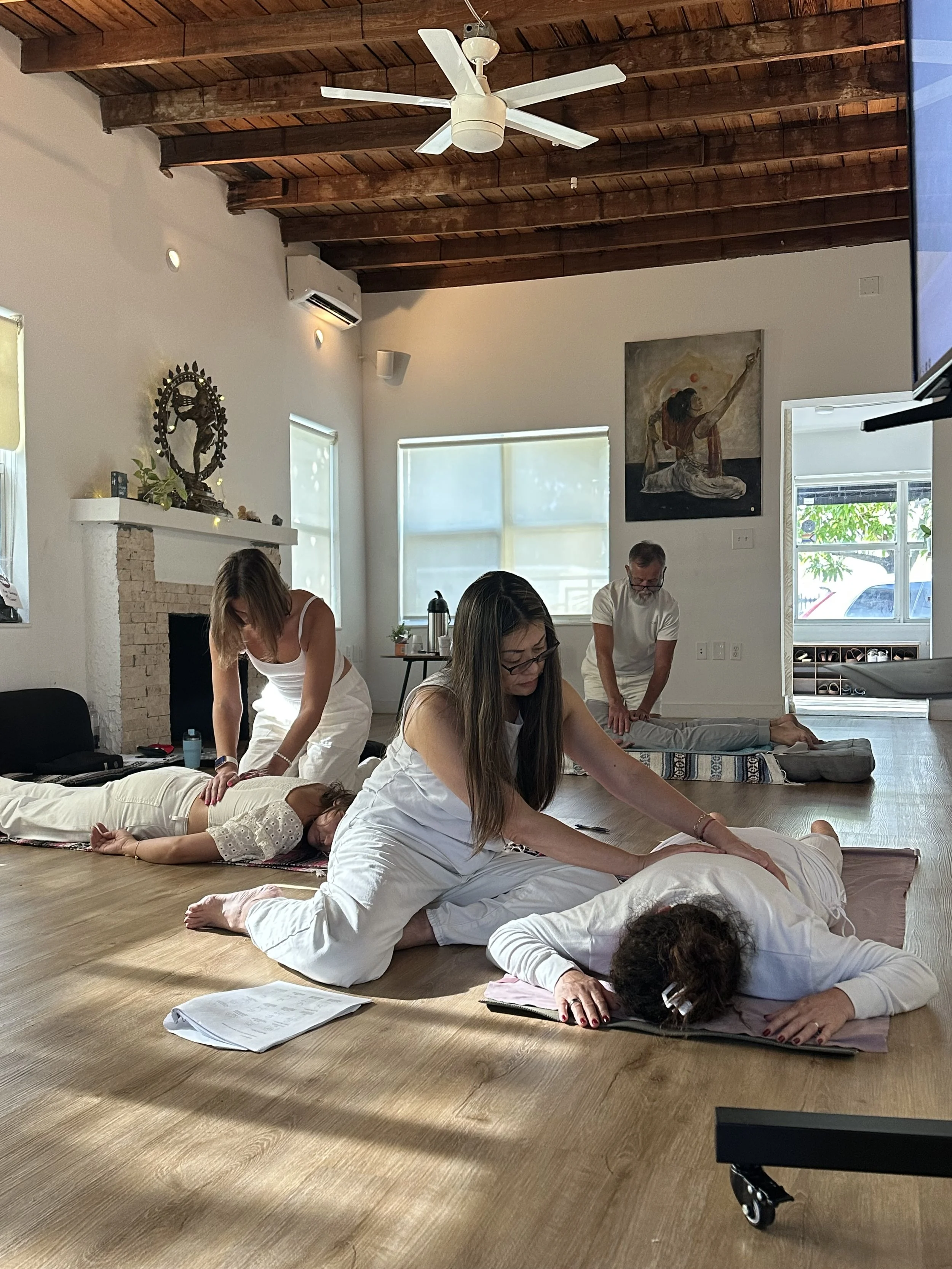 People practicing yoga or a similar stretching activity on mats in a well-lit room with wooden floors and a wooden ceiling, with a ceiling fan and artwork on the walls.