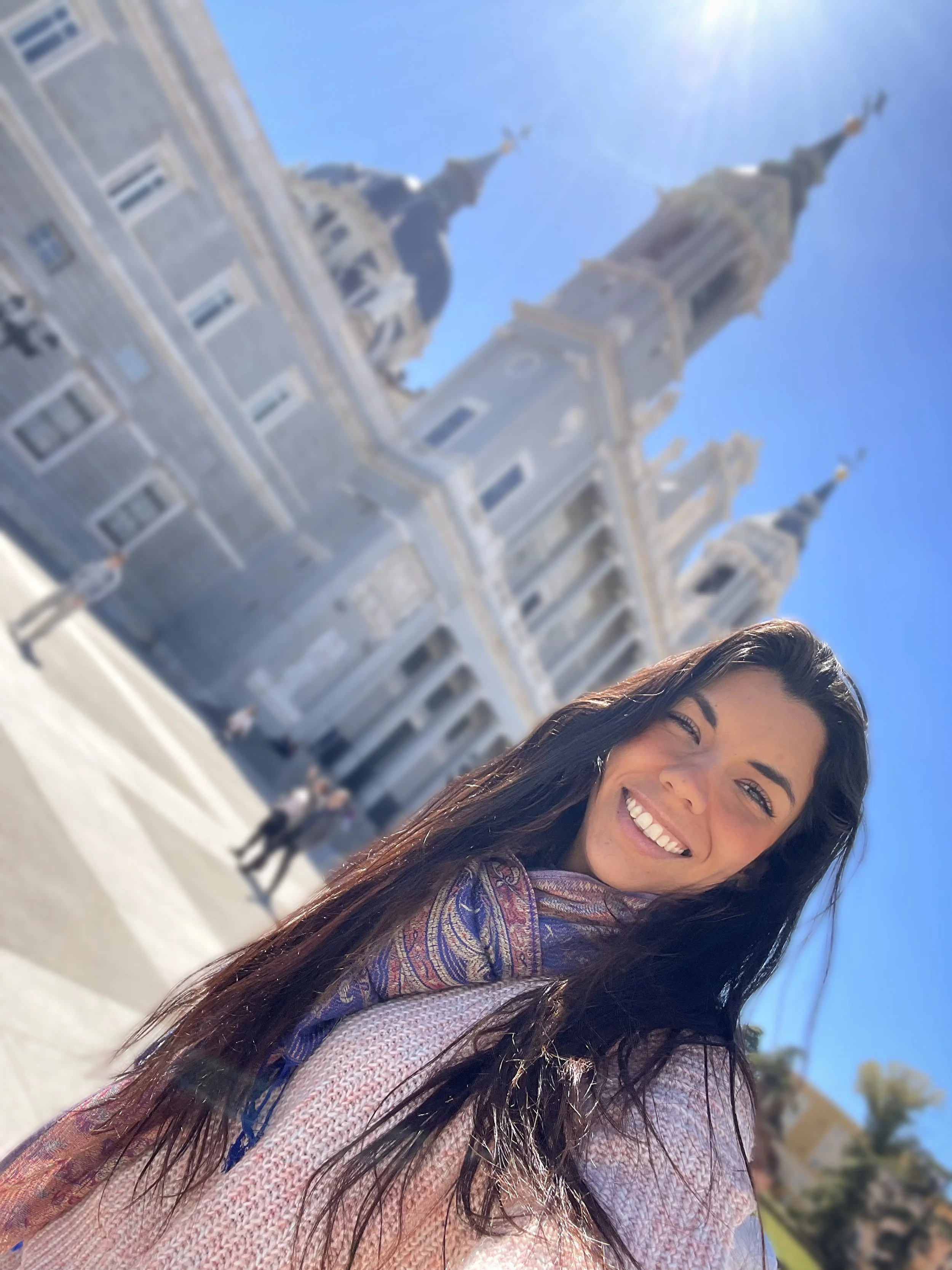 A smiling woman with long dark hair taking a selfie in front of a historic church with tall spires and a clear blue sky.