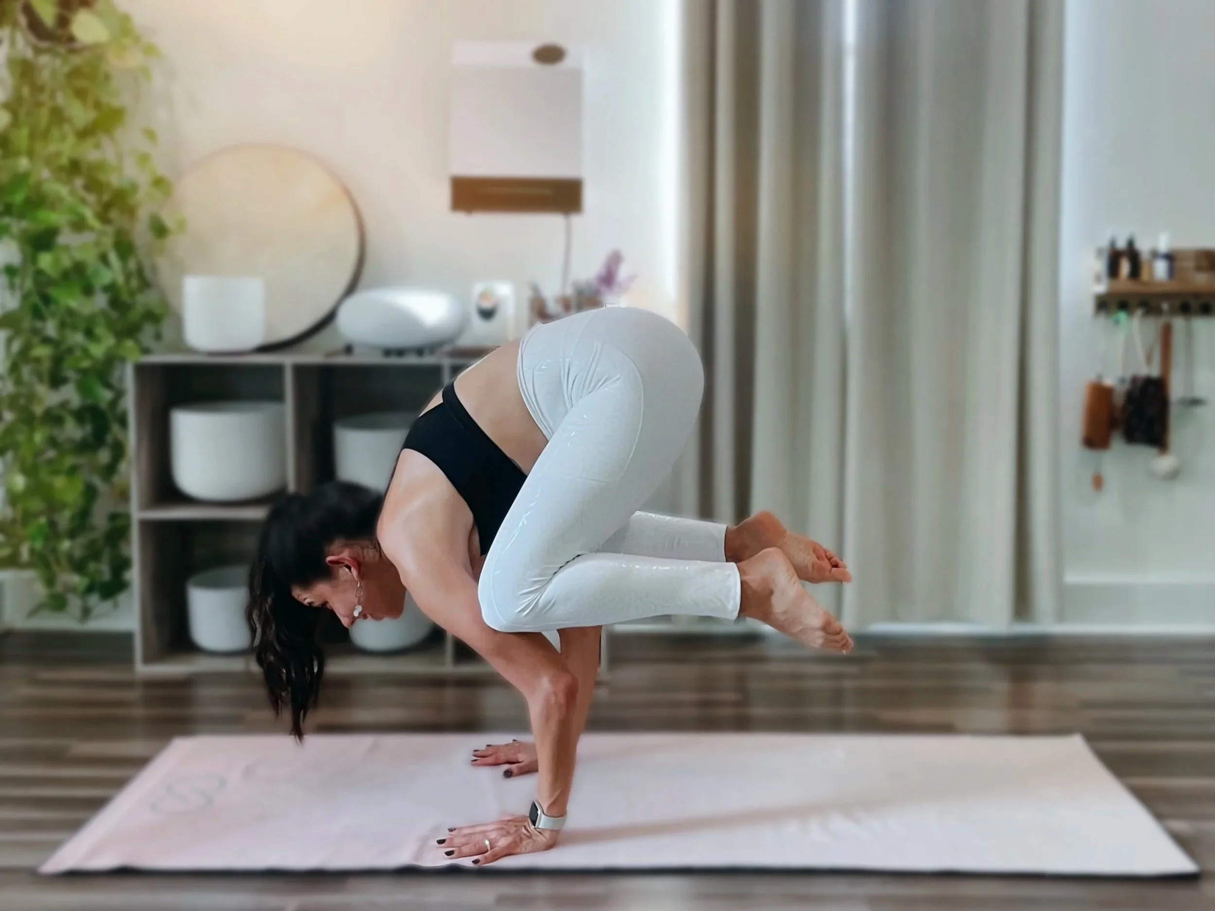 A woman practicing yoga in a bright, modern studio, balancing on one leg with her hands on the floor, wearing white leggings and a black top.