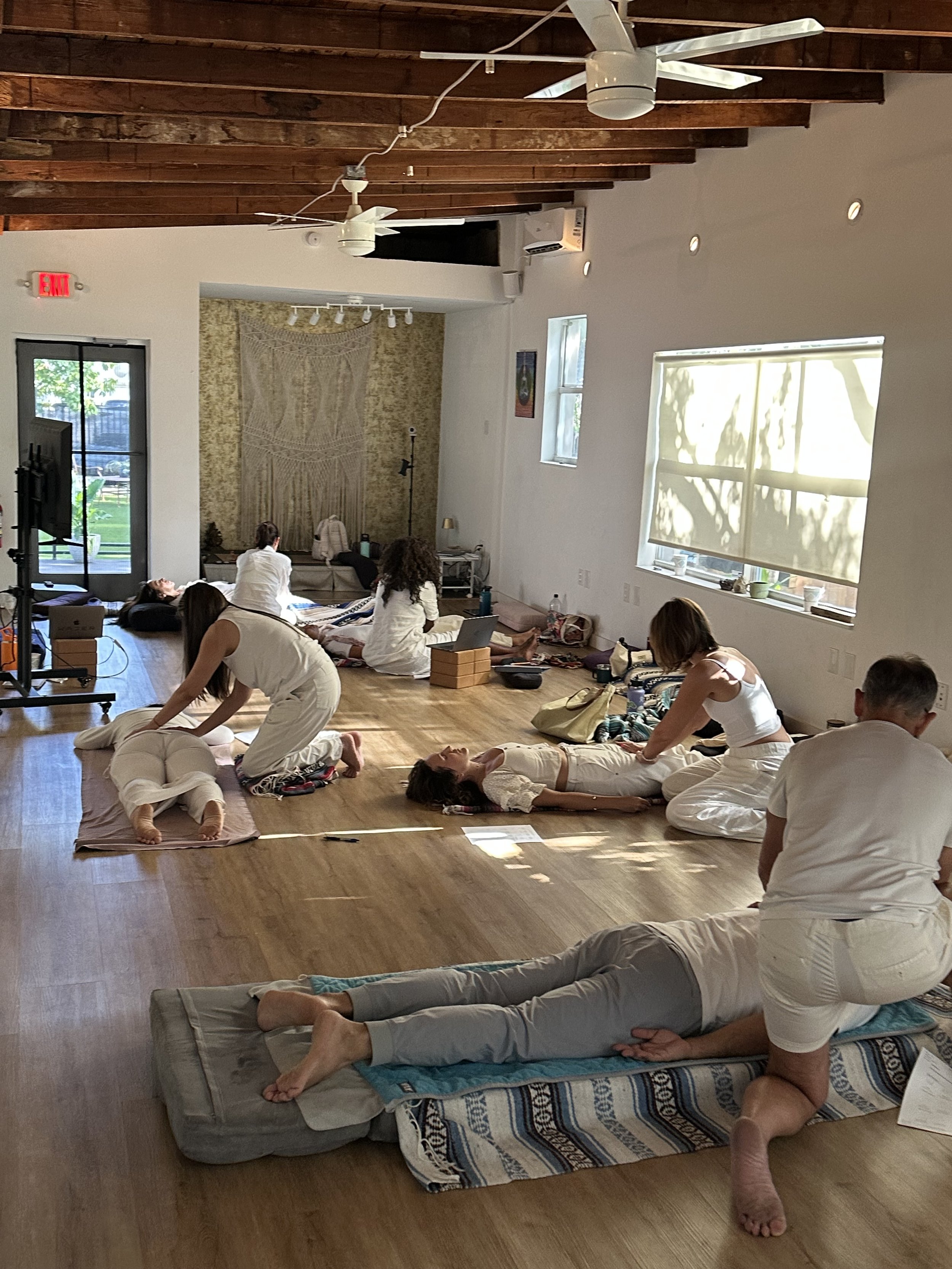 People practicing yoga or meditation in a bright, spacious room with wooden floors, large windows, and green outdoor scenery.
