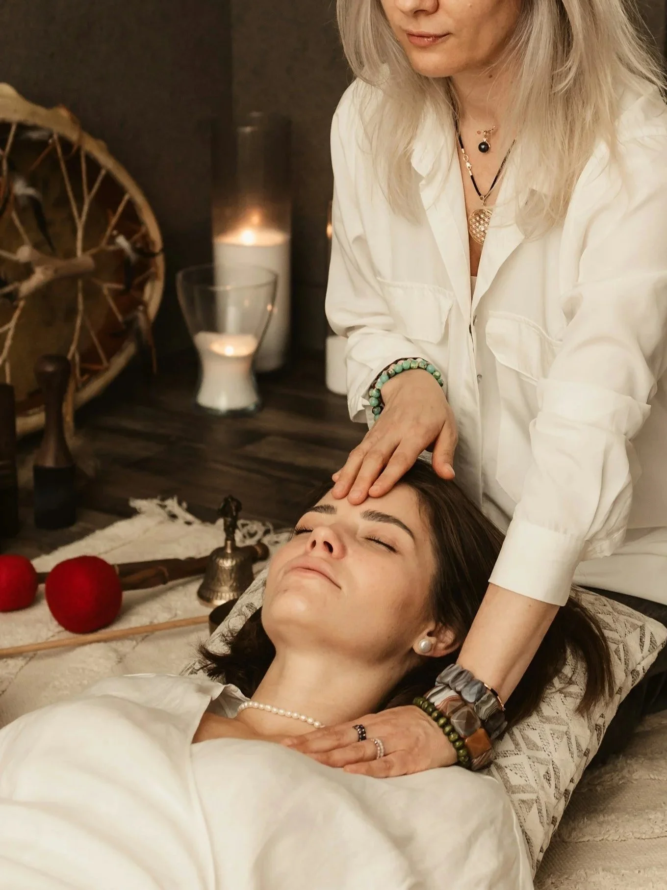 A person lying down with closed eyes receiving a massage or energy treatment from another person, who is gently placing their hand on the person's head. The scene is cozy with candles, decorative items, and red apples on a nearby surface.