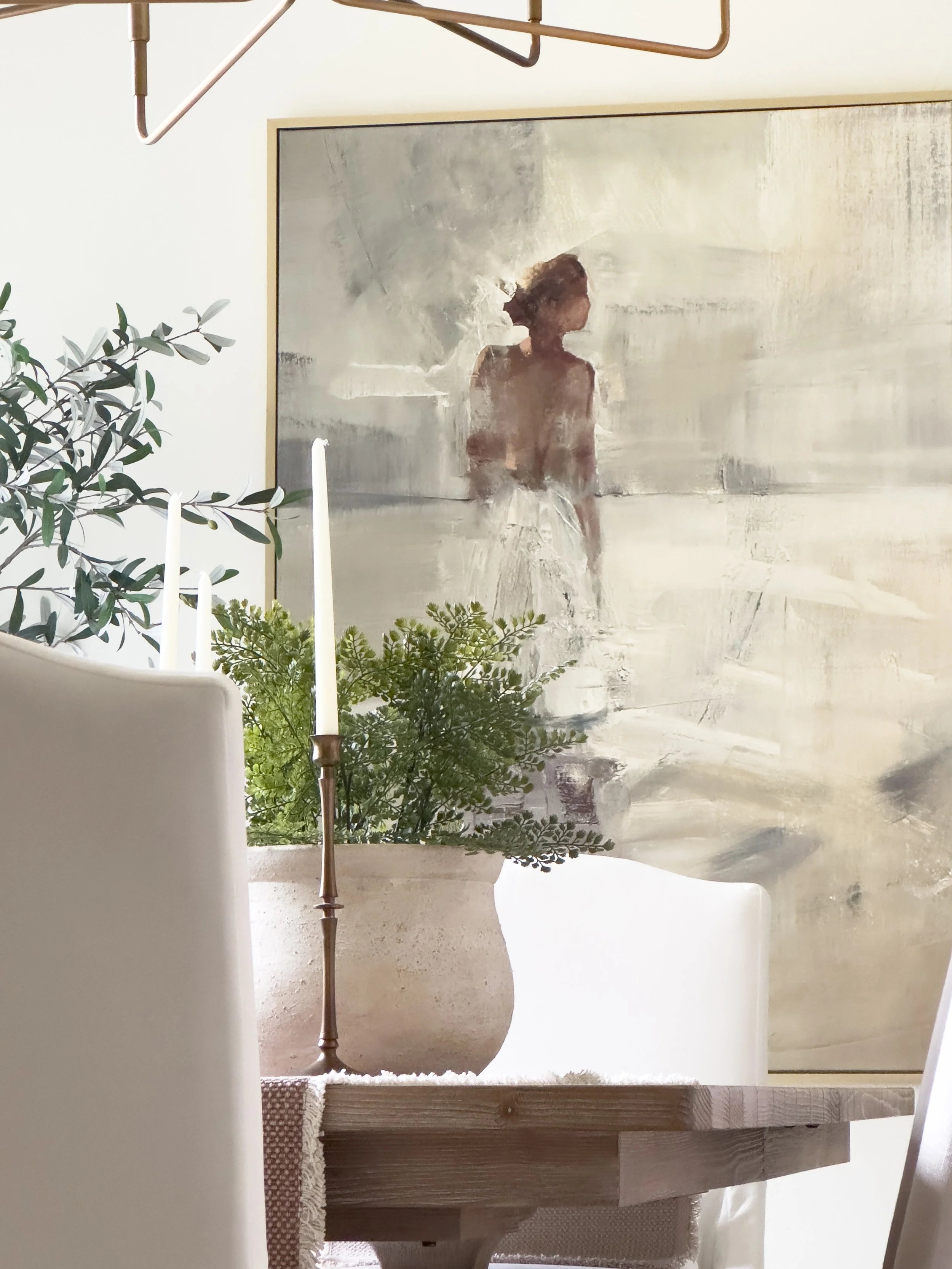 A decorative interior scene with a potted green plant, a candle holder with a tall white candle, and a framed abstract painting of a girl with curly hair, all arranged on a table with a textured beige table runner. The scene appears cozy and artistic with neutral tones.
