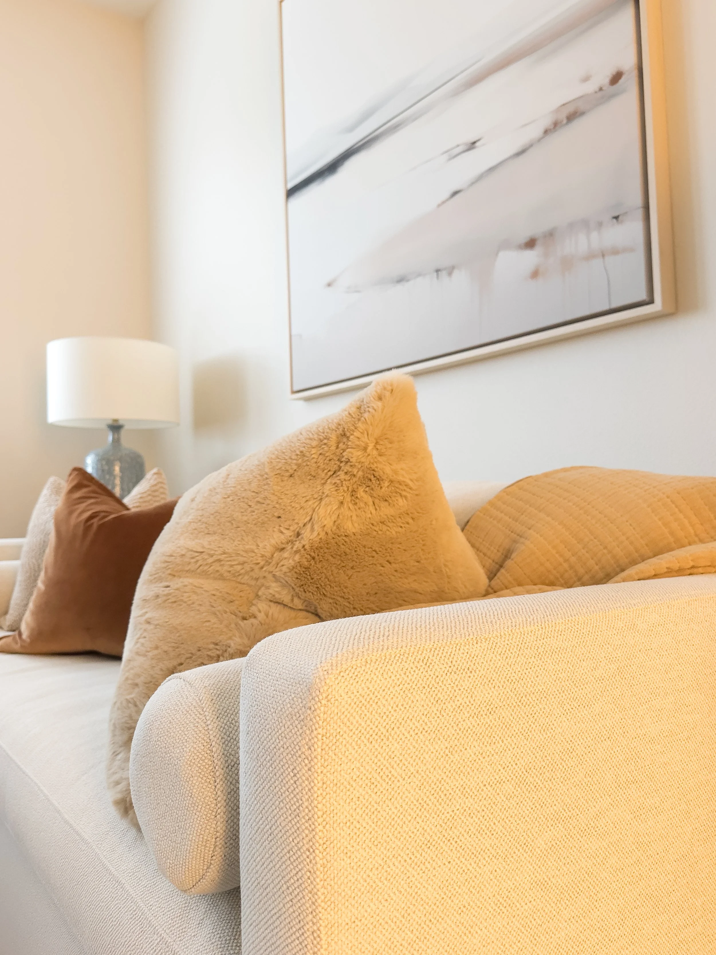 Close-up of a light-colored sofa with various textured cushions, in a room with a lamp and a large abstract painting on the wall.