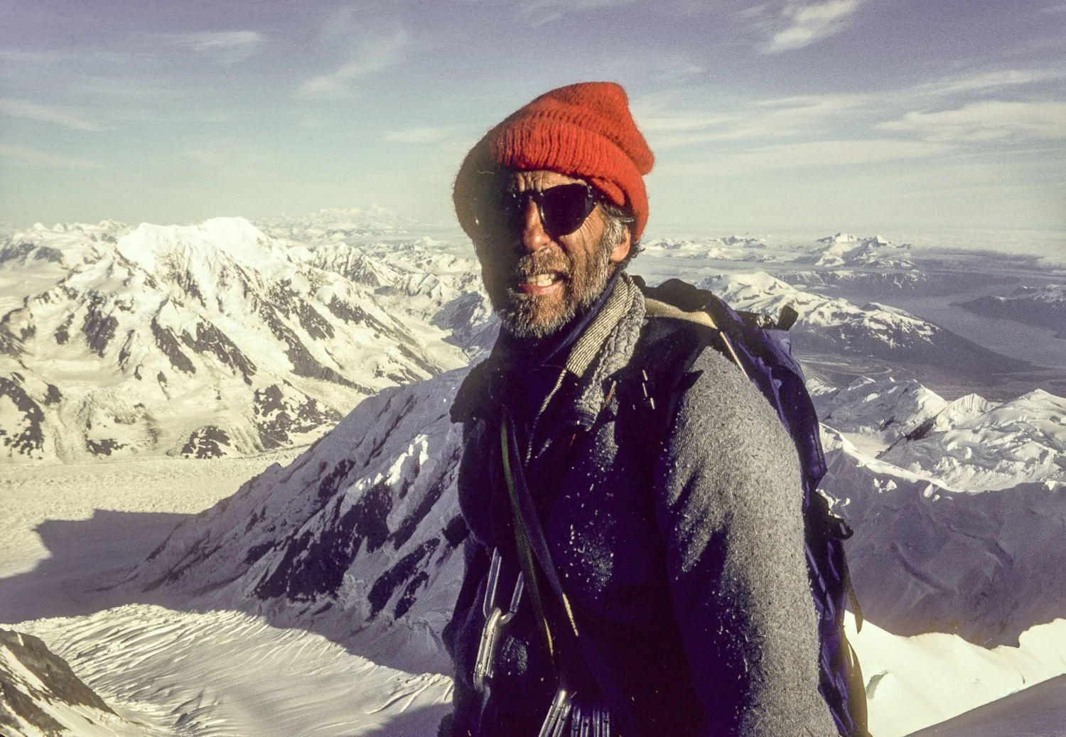 Mountaineer wearing a red hat and sunglasses stands on a snowy summit with expansive mountain range in the background
