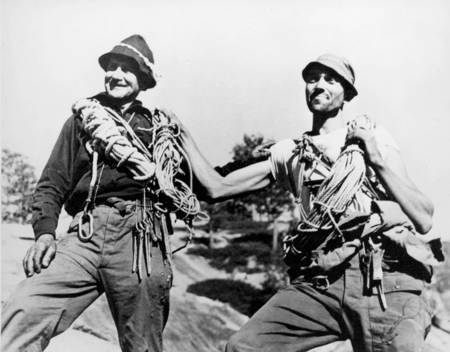 Two climbers stand side by side smiling, wearing hats and carrying coils of rope and climbing gear outdoors