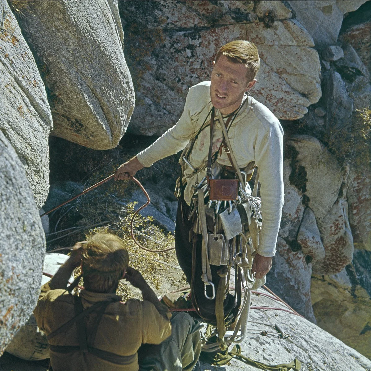 Climber in Yosemite stands on a ledge with gear, looking up while belaying partner seated below on rocky wall