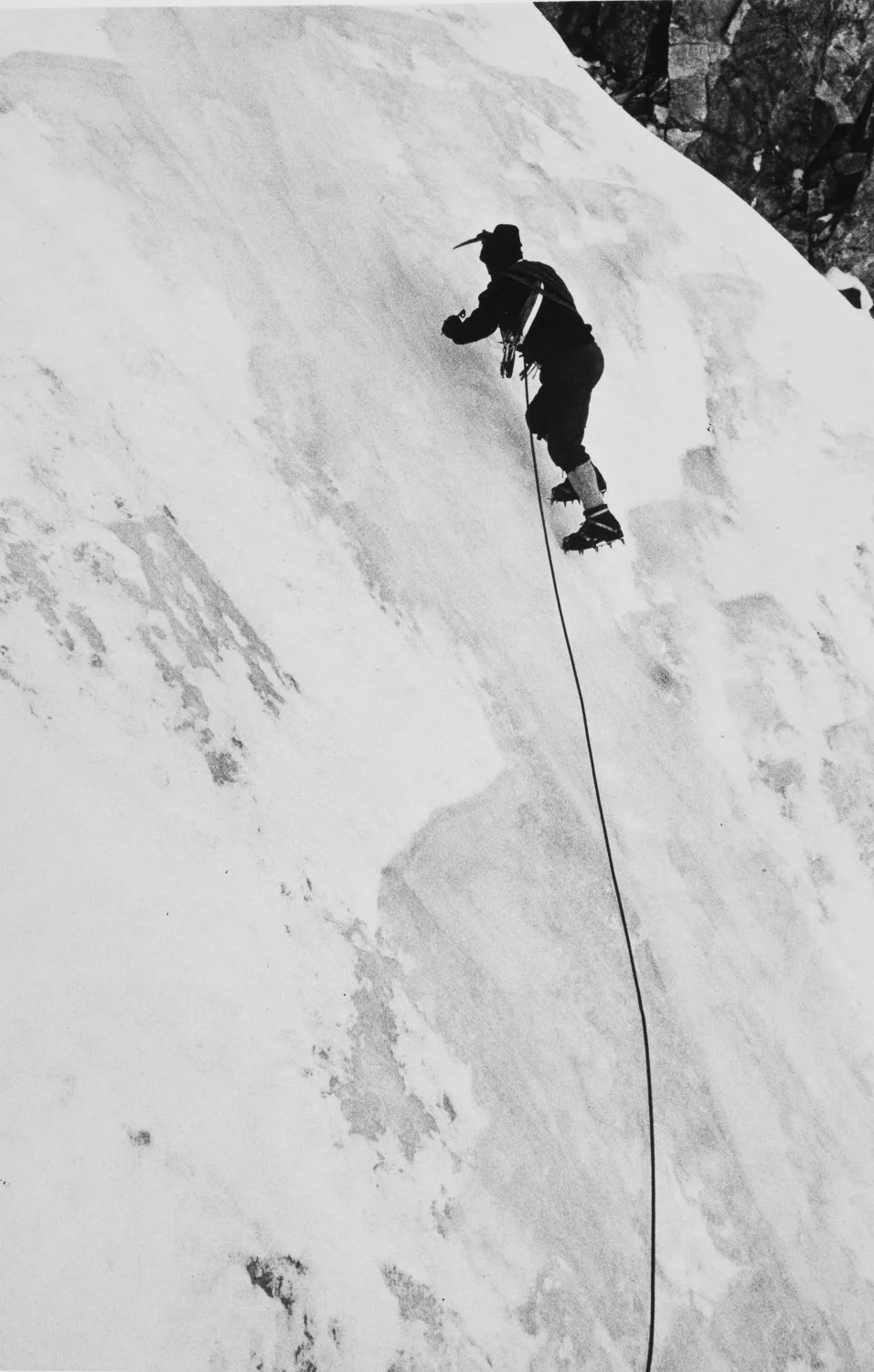 Climber ascends a steep icy face on rope, using crampons and ice tools on a near-vertical snow-covered wall