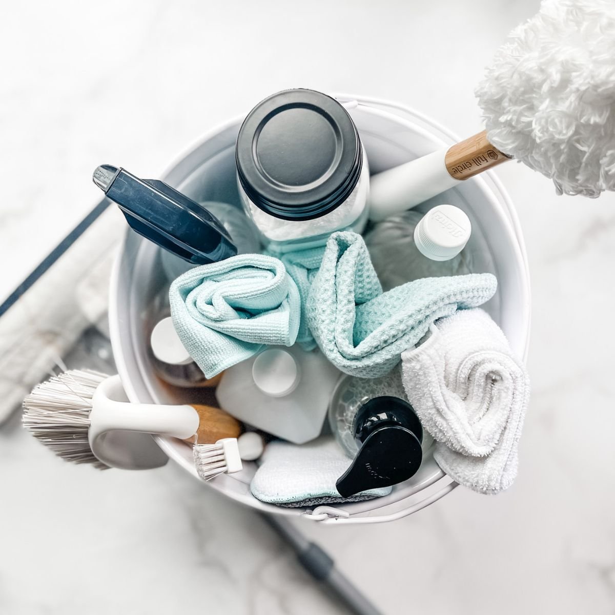 A top-down view of a white container filled with cleaning supplies, cloths, spray bottles, and a scrub brush.