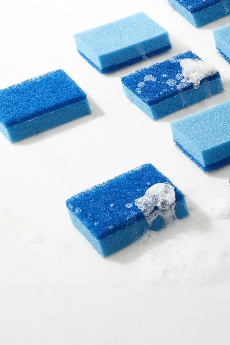 Blue and light blue cleaning sponges on a white surface, some are covered with soap suds and foam.