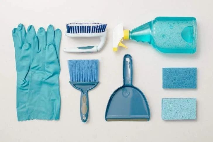 Cleaning supplies including blue rubber gloves, scrub brushes, a dustpan, a spray bottle, and cleaning sponges arranged on a white surface.