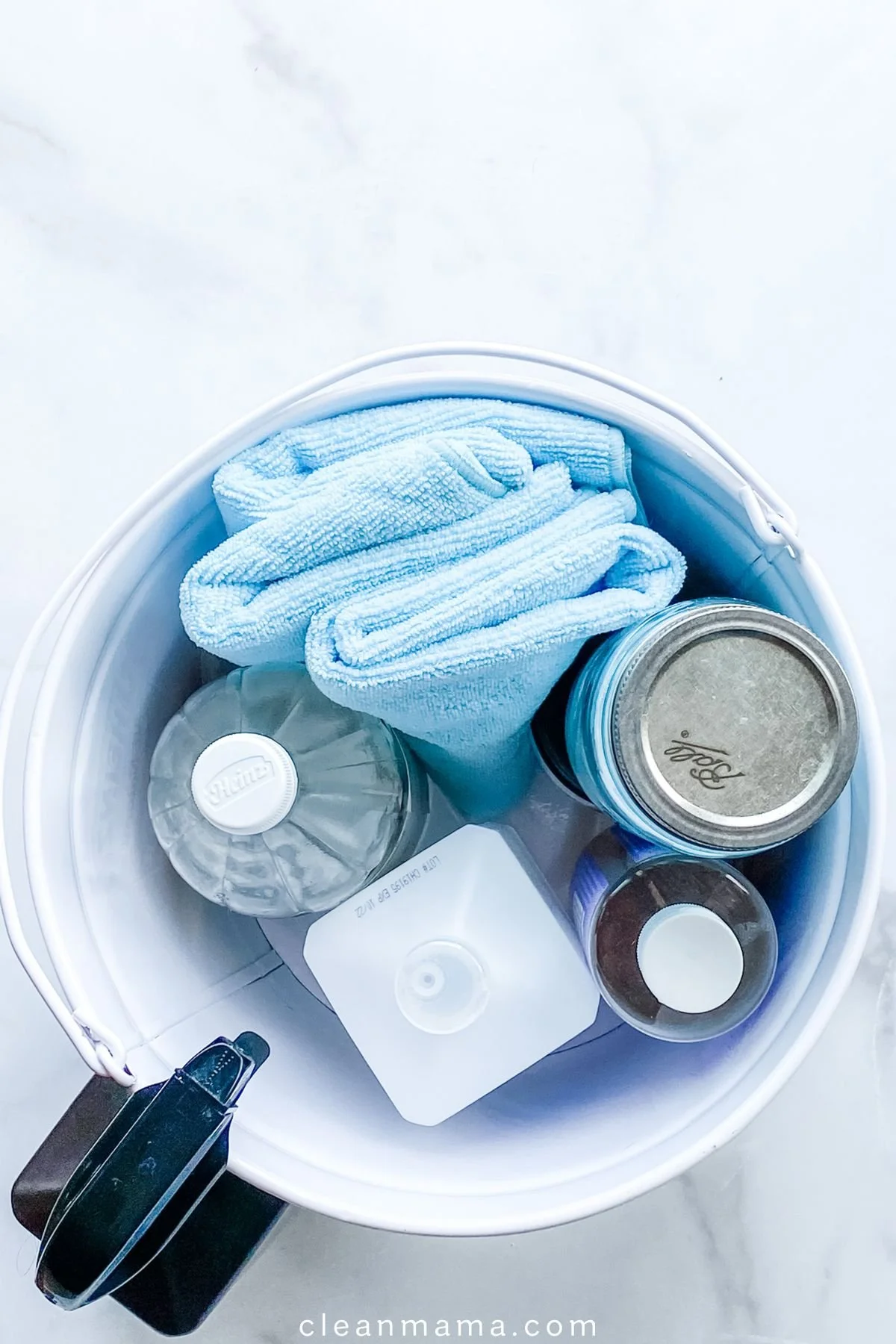 A white bucket filled with cleaning supplies including a blue microfiber cloth, a clear water spray bottle, a container with a metal lid, and other cleaning bottles.