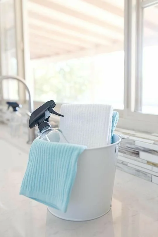 Cleaning supplies including spray bottle, cleaning cloths, and towels in a metal bucket on a kitchen countertop near a window.
