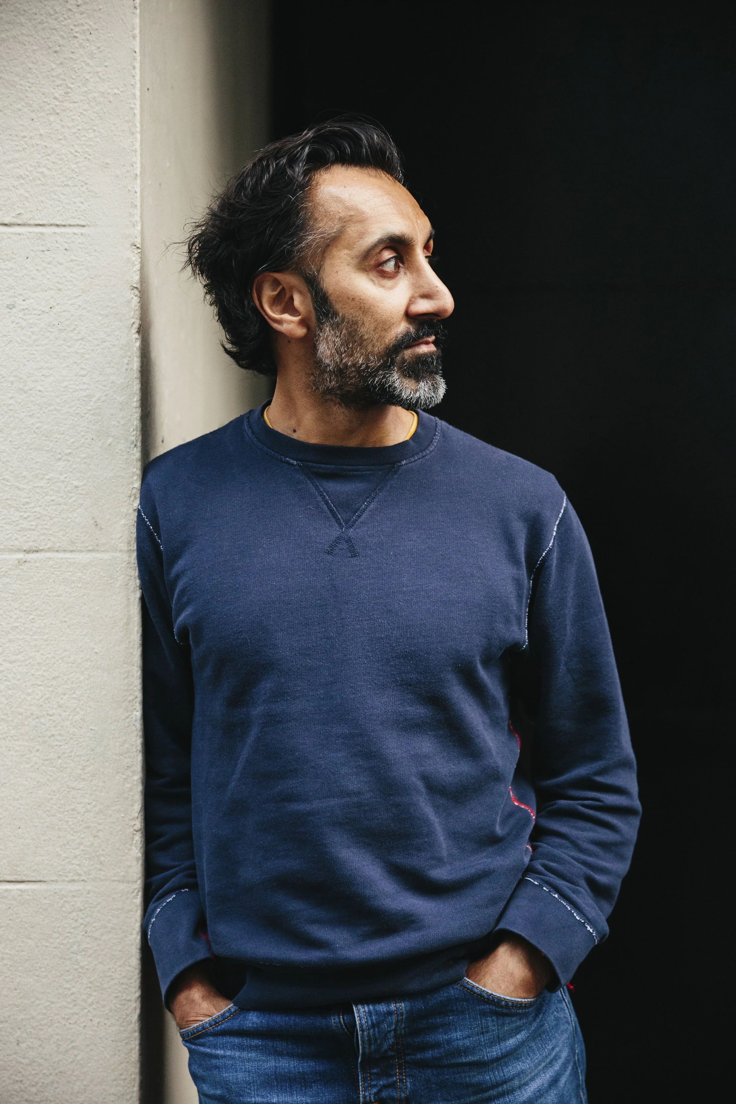 A man with dark hair and a beard, wearing a navy blue sweatshirt and jeans, leaning against a wall in front of a black background, looking to his left.