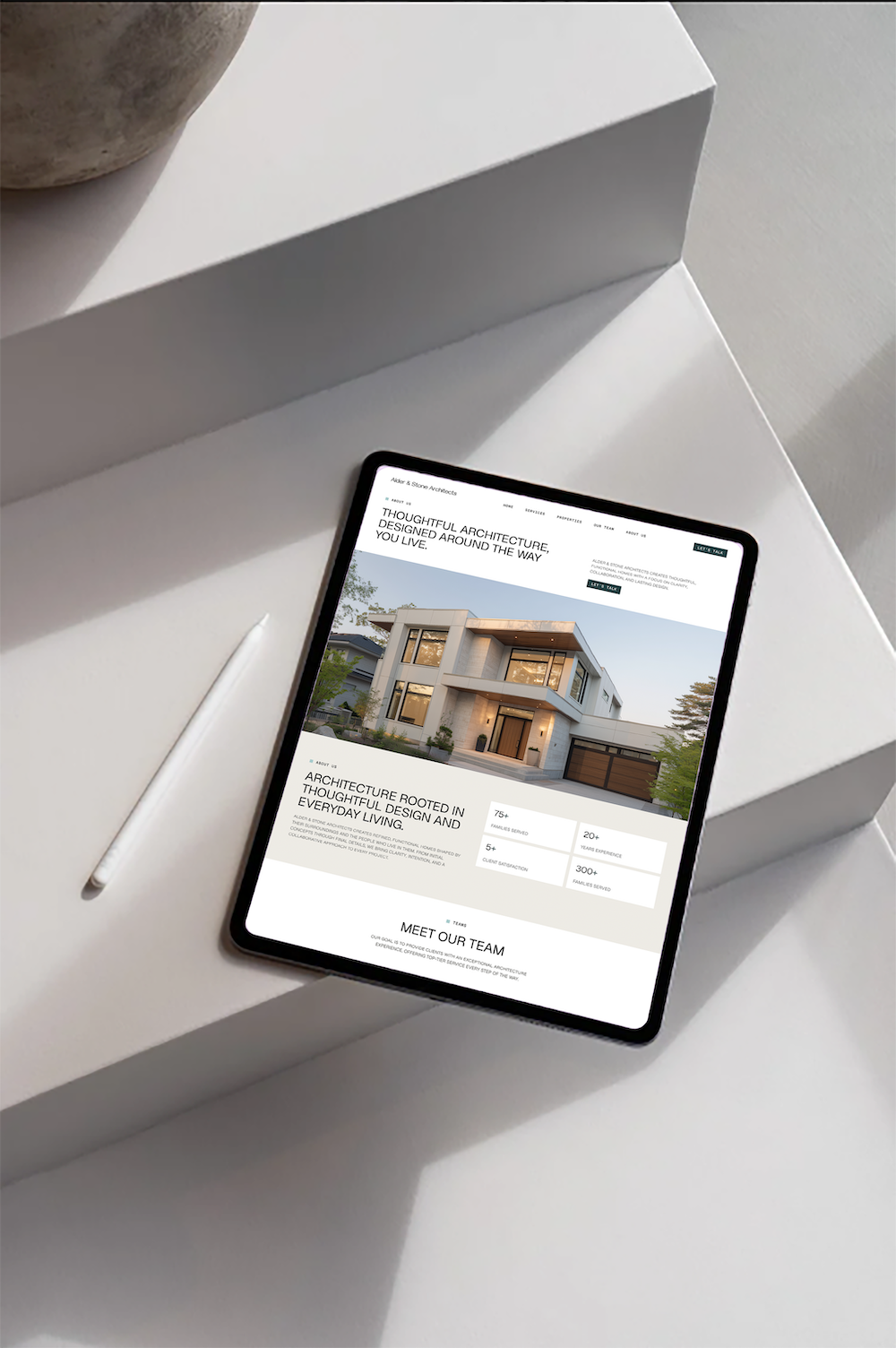 An iPad displaying a website for architecture firm with a modern house image, placed on a white table next to a white rectangular box and a gray decorative object.