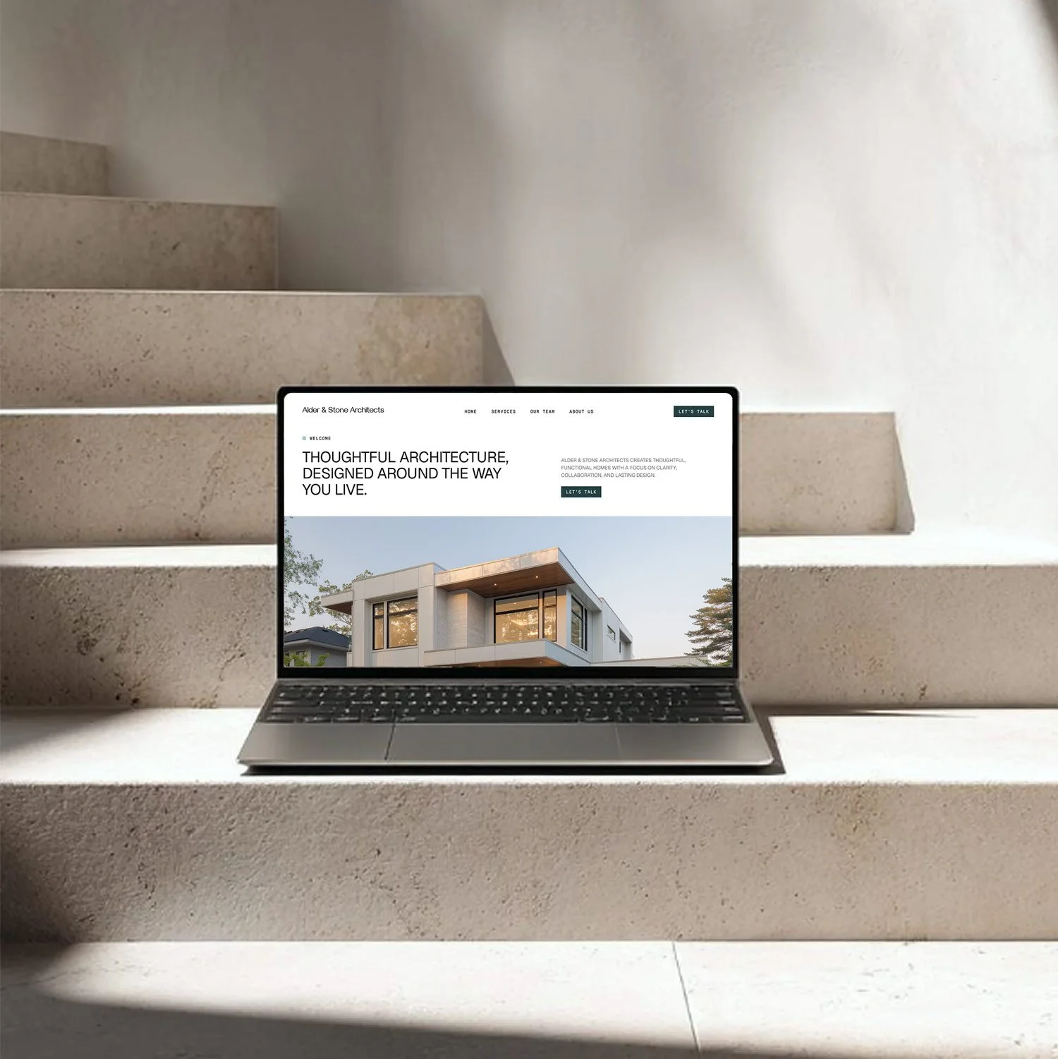 Laptop displaying a website for Alder & Stone Architects, featuring a modern house design, placed on a concrete staircase with sunlight casting shadows.