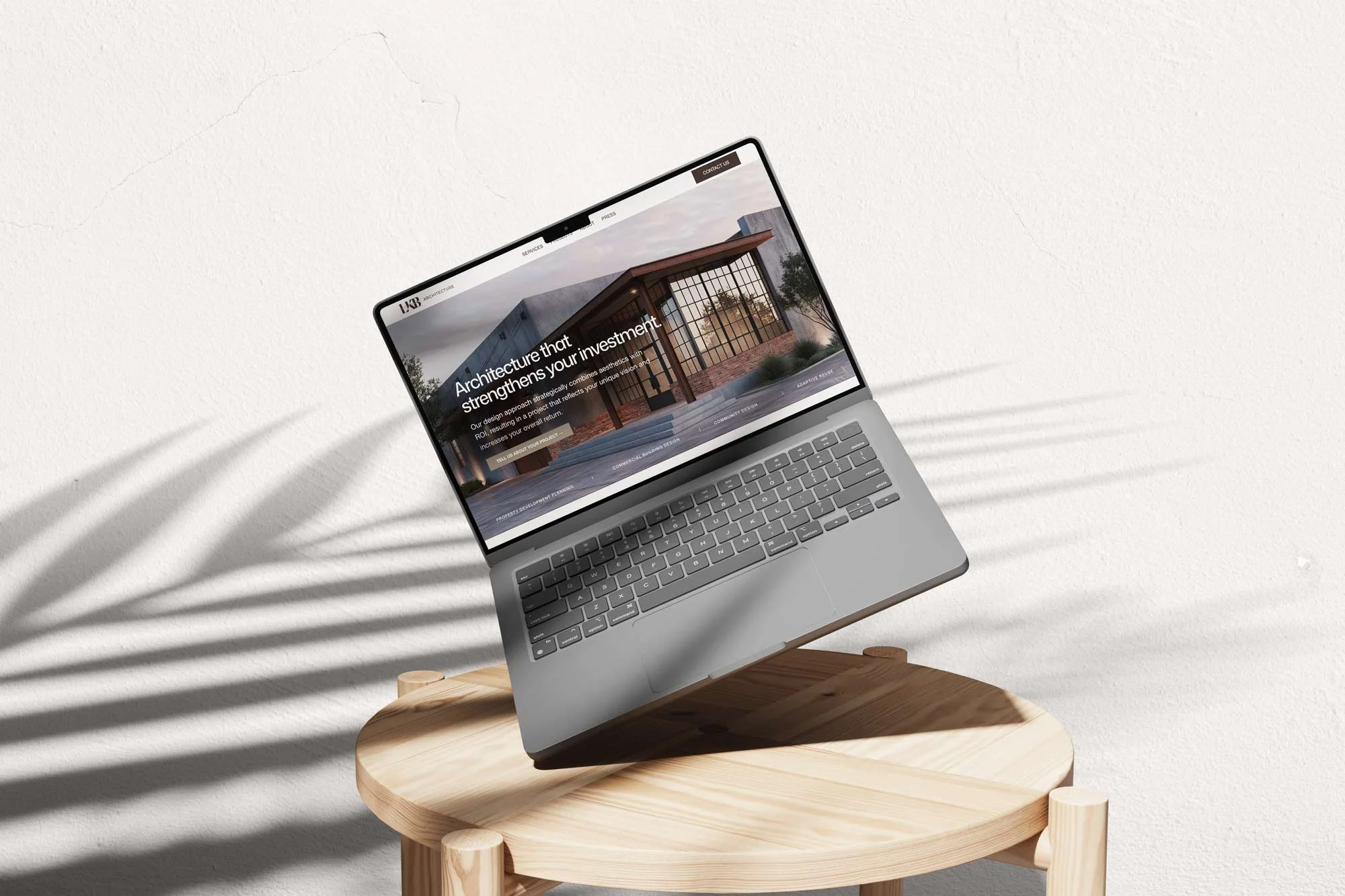 A silver laptop on a small round wooden table displaying a webpage about architecture projects, with text about strengthening investments through architecture.