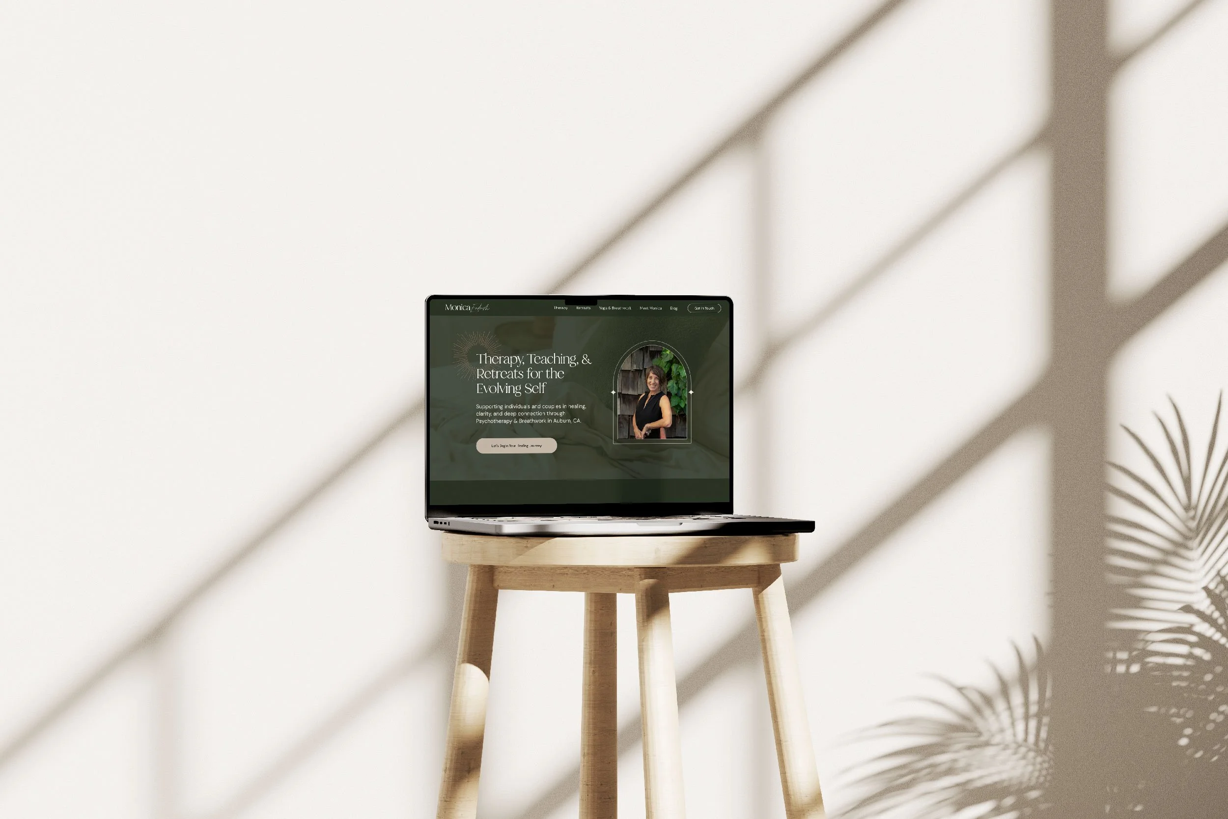 Laptop displaying a webpage about therapy, teaching, and retreats for the evolving self, placed on a wooden stool against a white wall with shadows and plant leaves partially visible.