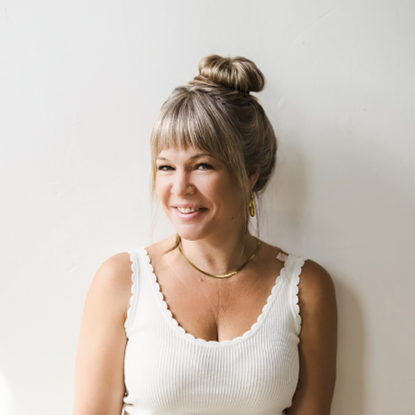 A woman with blonde hair in a bun, wearing a white sleeveless top, smiling and standing against a plain white wall.