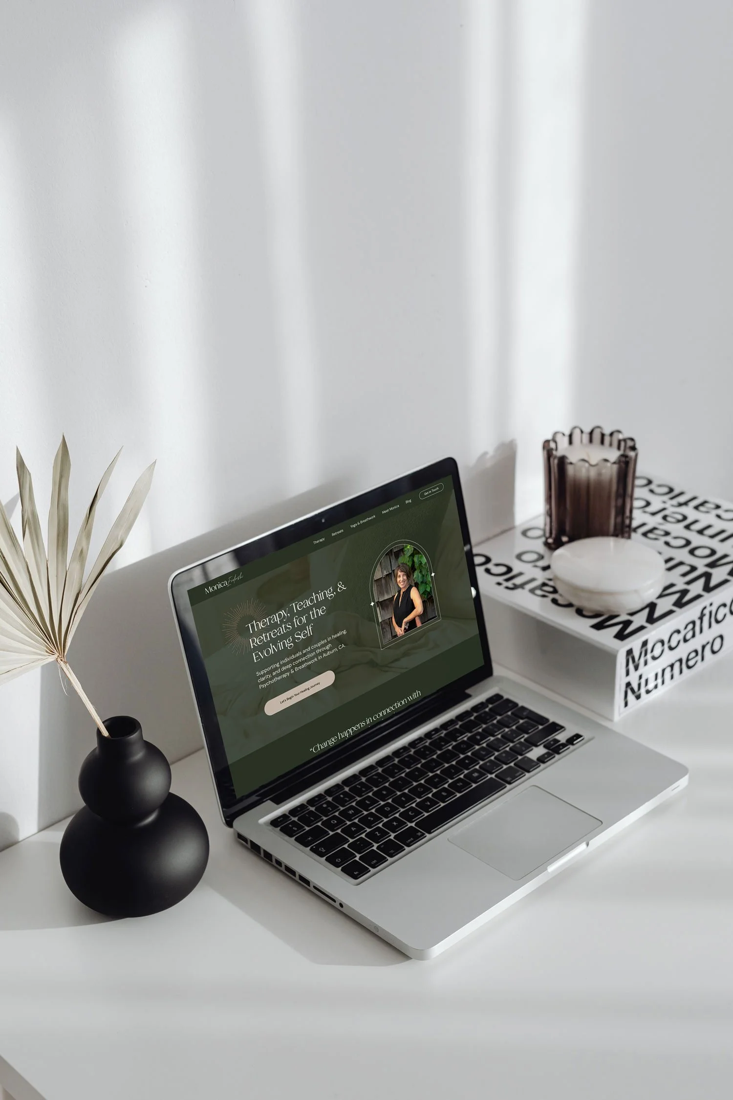 A workspace with a silver laptop displaying a website about therapy and retreats, a black vase with dried leaves, a decorative dark brown chair, a white round container, and a box with black and white text on a white surface in front of a white wall.