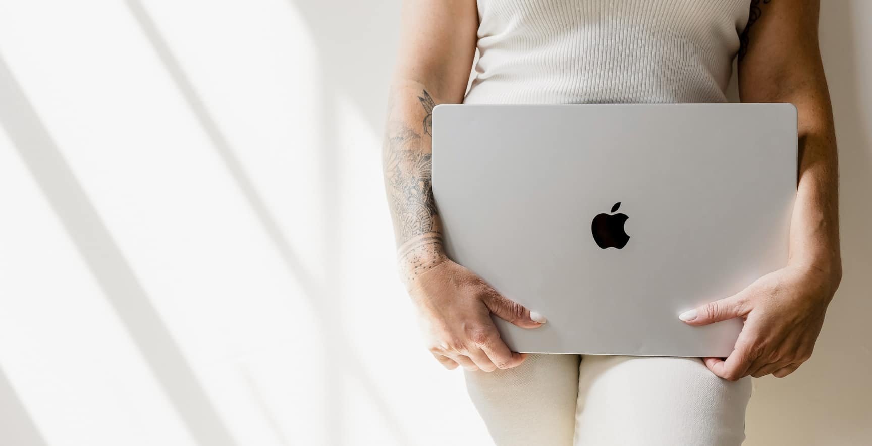 Person holding a silver MacBook laptop with an Apple logo, wearing a white sleeveless top, with tattoos on one arm, standing against a white wall.