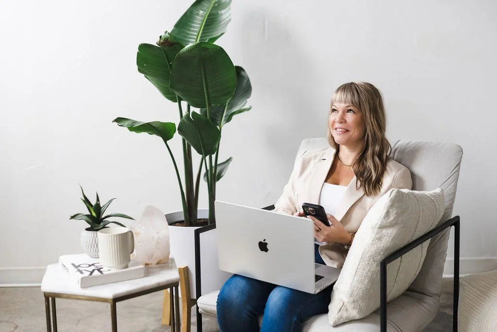 A woman sitting on a beige chair with a laptop on her lap, smiling and holding a smartphone, in a minimalistic room with large green potted plants and a small side table with a smaller plant, salt lamp, and books.