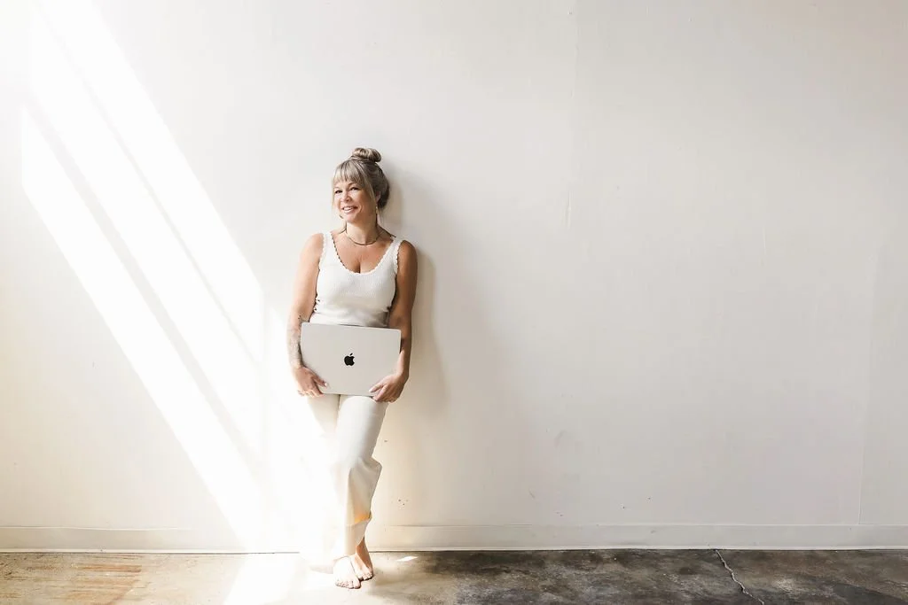 A woman with gray hair in a bun, wearing a white sleeveless top and pants, standing barefoot against a white wall with sunlight streaming in, holding a silver laptop.