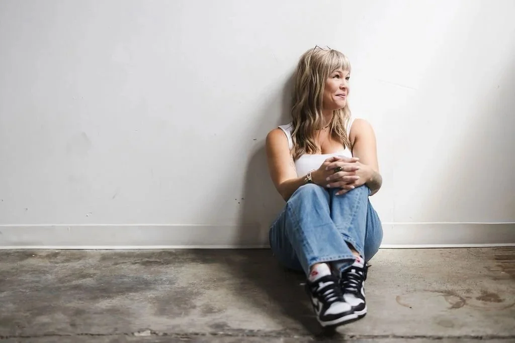 A woman with blonde hair sitting on the floor against a plain white wall, wearing a white tank top, blue jeans, and sneakers, looking to the right with a thoughtful expression.
