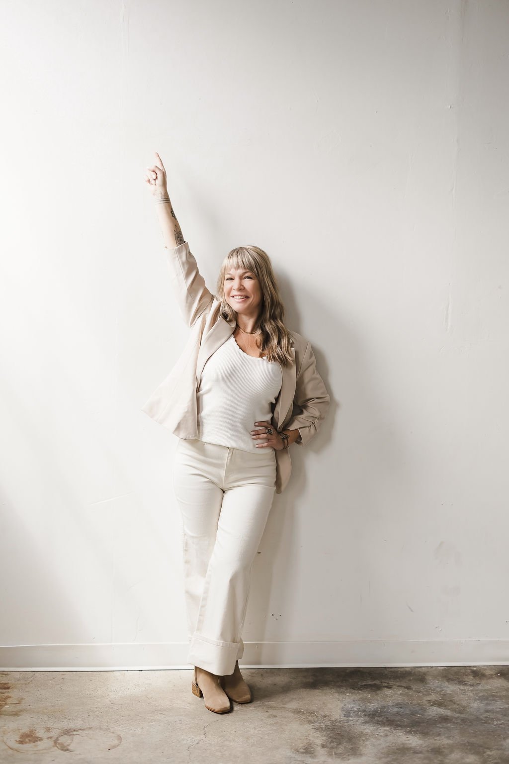 Woman with long curly hair wearing a beige blazer and pants, smiling and raising her right arm.