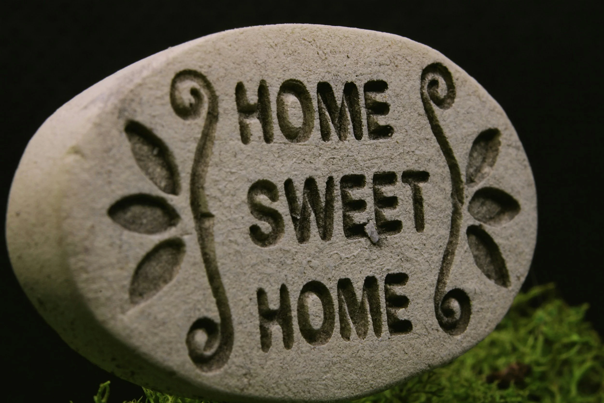 Close-up of a carved stone with the words 'HOME SWEET HOME' and decorative hearts on it, placed on a patch of grass.