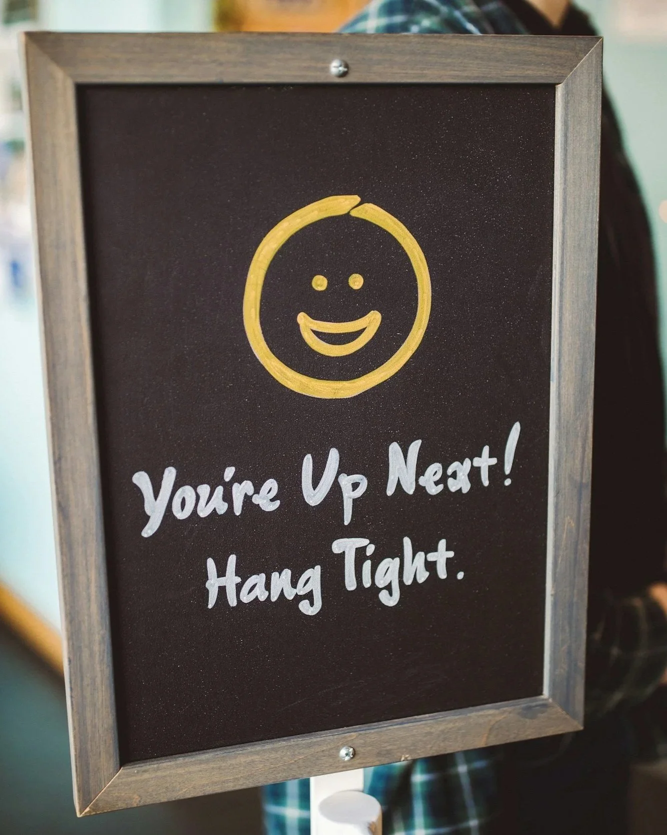 A blackboard sign with a yellow smiley face at the top, and handwritten white text that says, "You're Up Next! Hang Tight."
