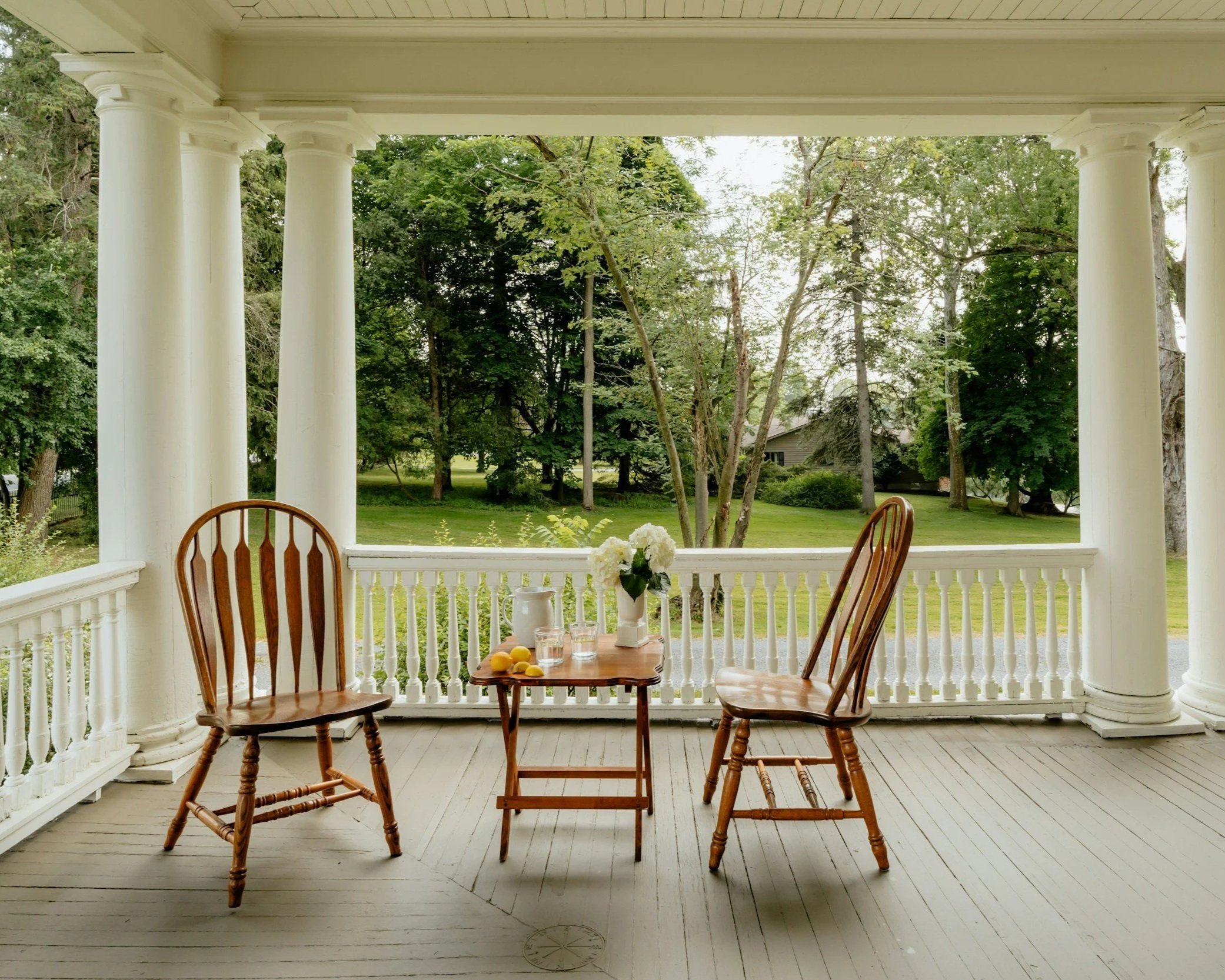 Porch with wooden chairs, a table with a vase of flowers, and trees in the background.