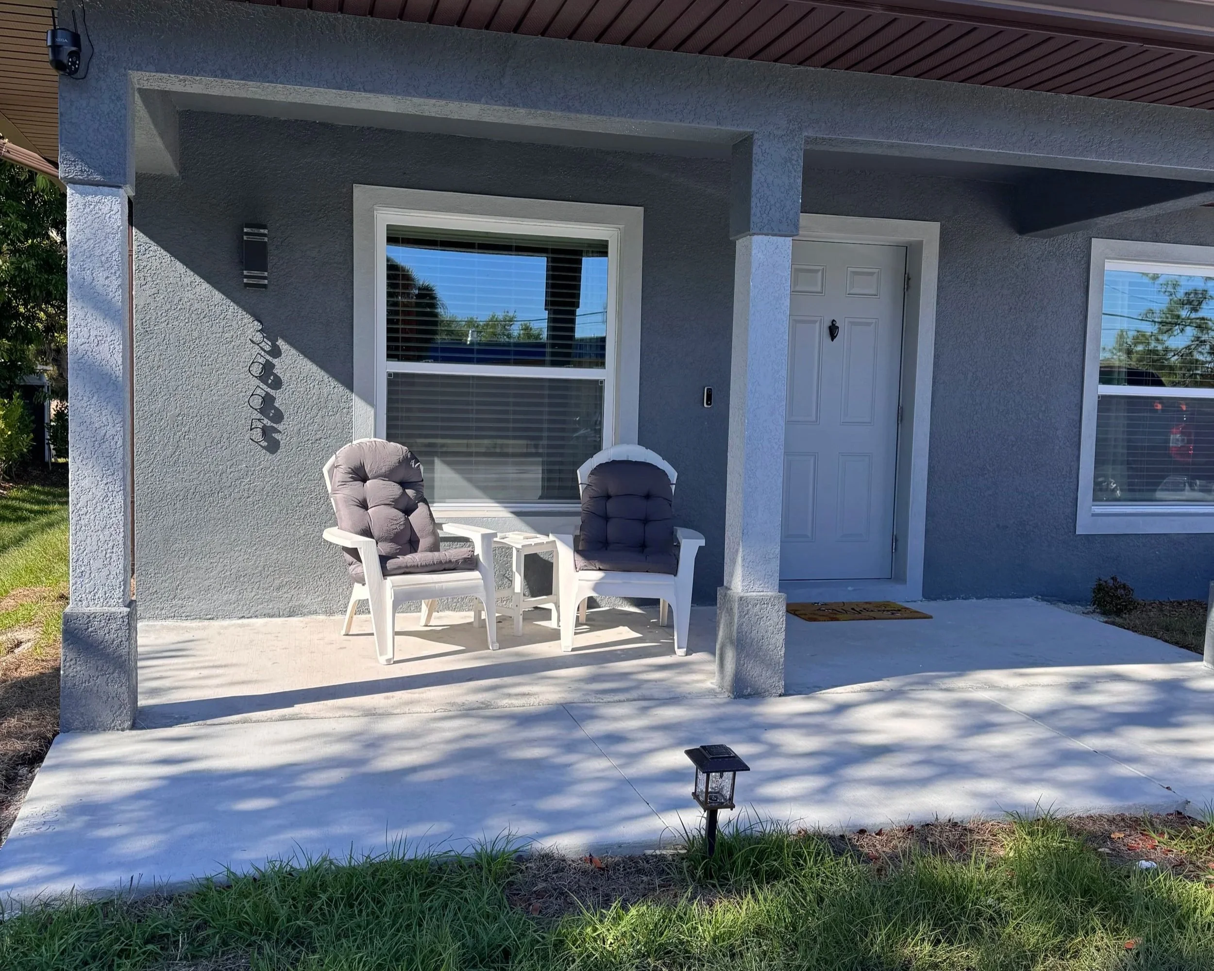 Backyard porch with two cushioned chairs, a white door, large windows, and a small outdoor lantern.