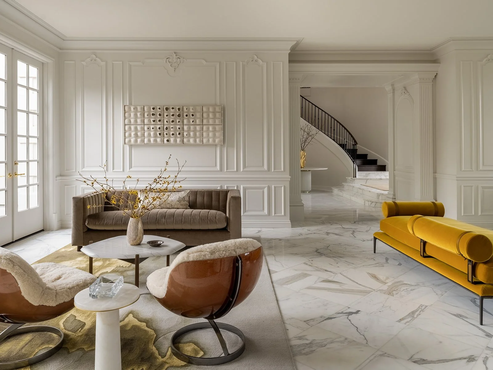 Elegant living room with white paneled walls, marble flooring, and contemporary furniture, including a beige sofa, brown and white chairs, a yellow velvet bench, and a white coffee table with a vase of branches. Rucon Build Work