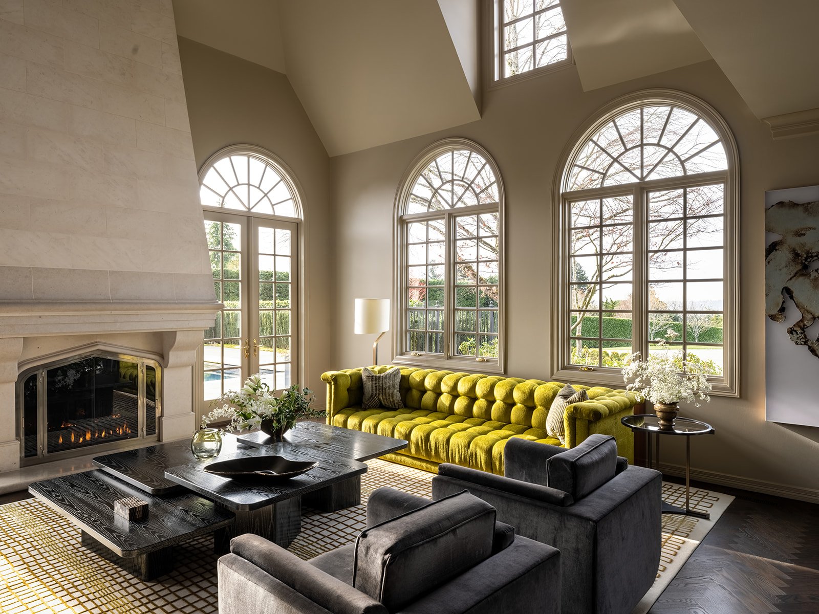 Living room with large arched windows, a yellow tufted sofa, a black coffee table, gray armchairs, a fireplace, and decorative plants.
