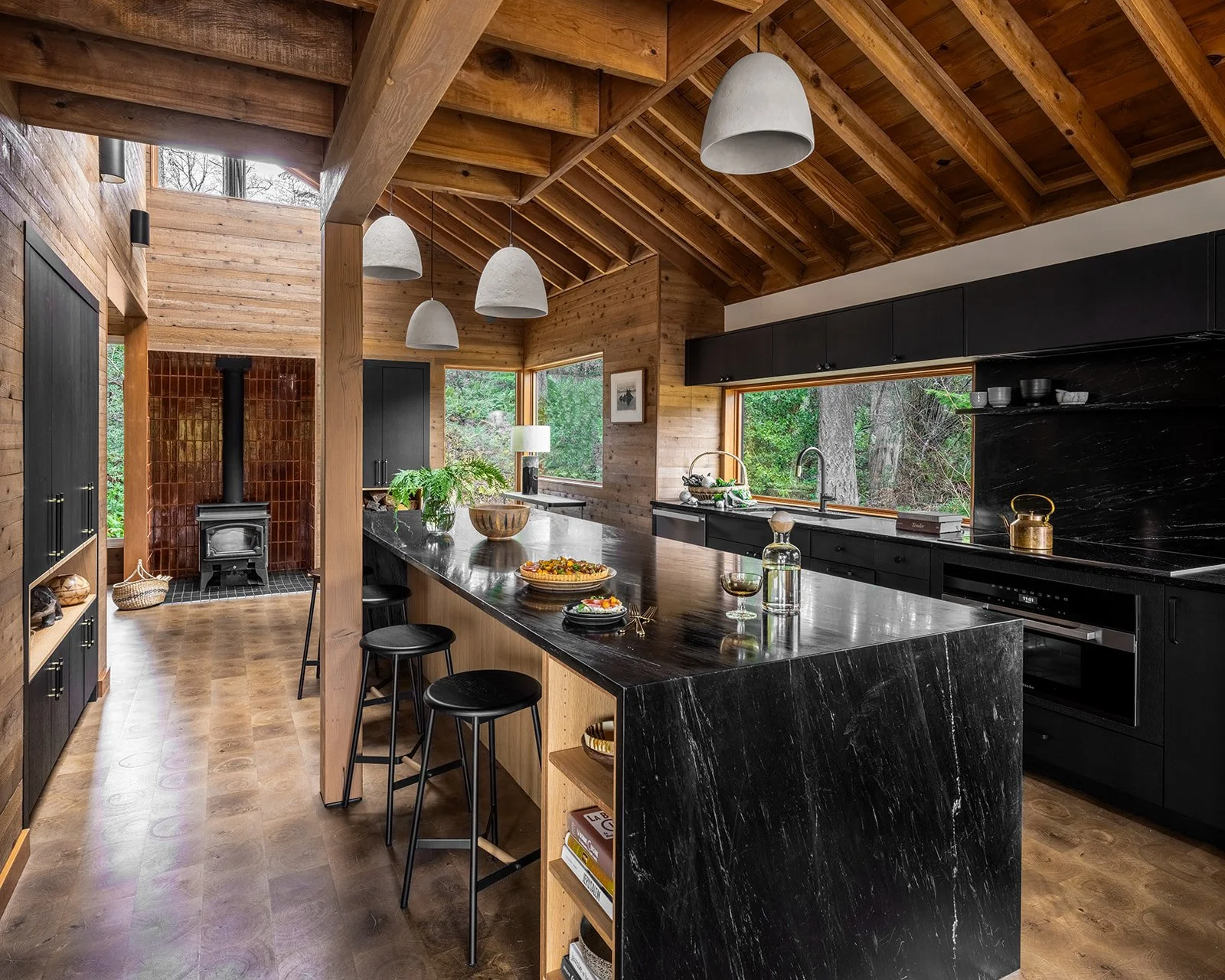 Interior of a modern kitchen with wooden walls and ceiling, black cabinets, a large black marble island with bar stools, gold accents, large windows showing greenery, and a wood-burning stove with a red tile back wall.